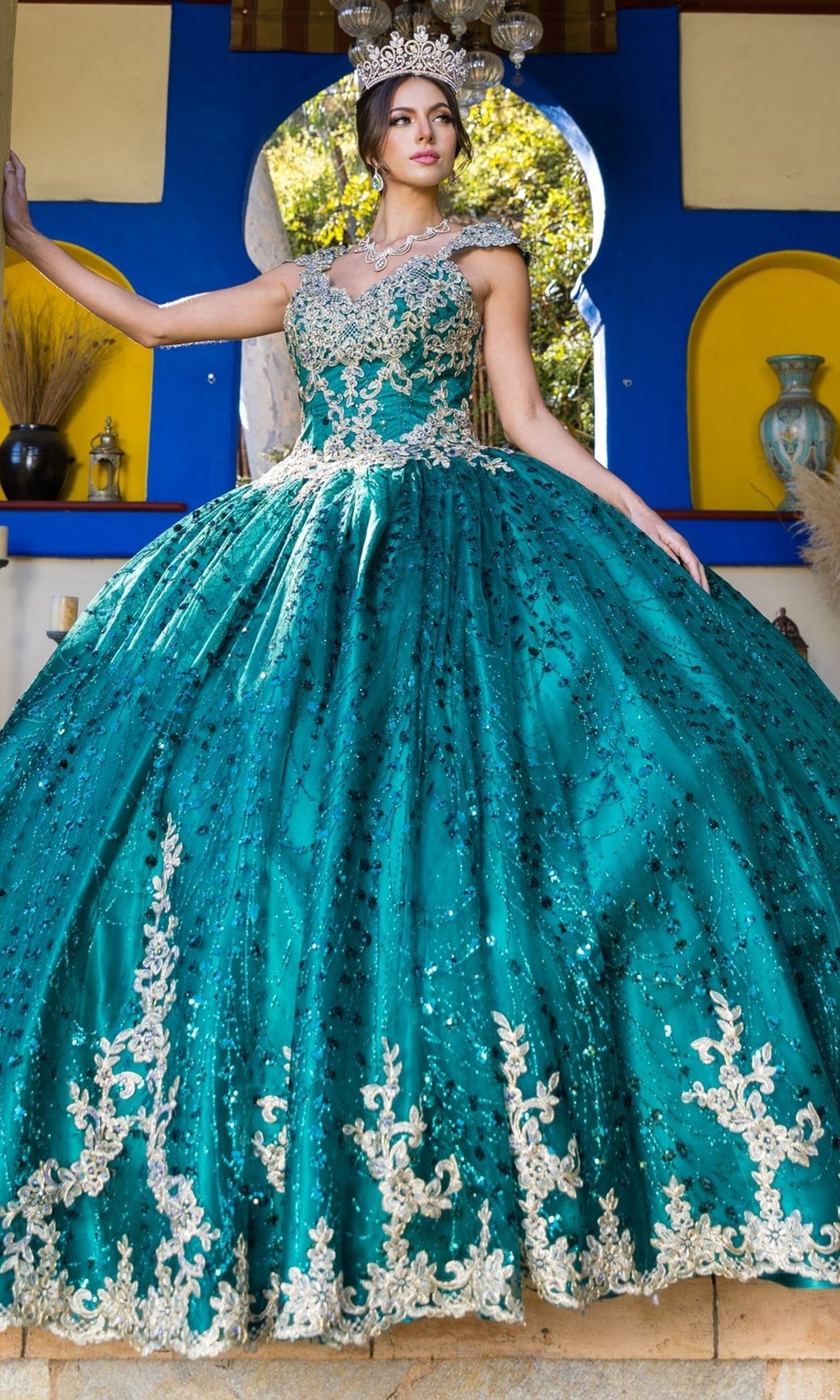 A woman in the Quinceanera Ball Gown 8024J, featuring gold lace embroidery and glitter tulle, stands before a vibrant background, wearing a jeweled crown.