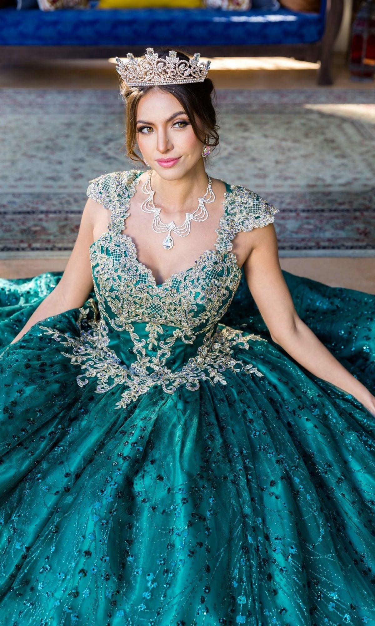 A woman in the Quinceanera Ball Gown 8024J, featuring teal fabric and gold lace embroidery, with a jeweled crown and silver necklace, sits on the floor looking at the camera.