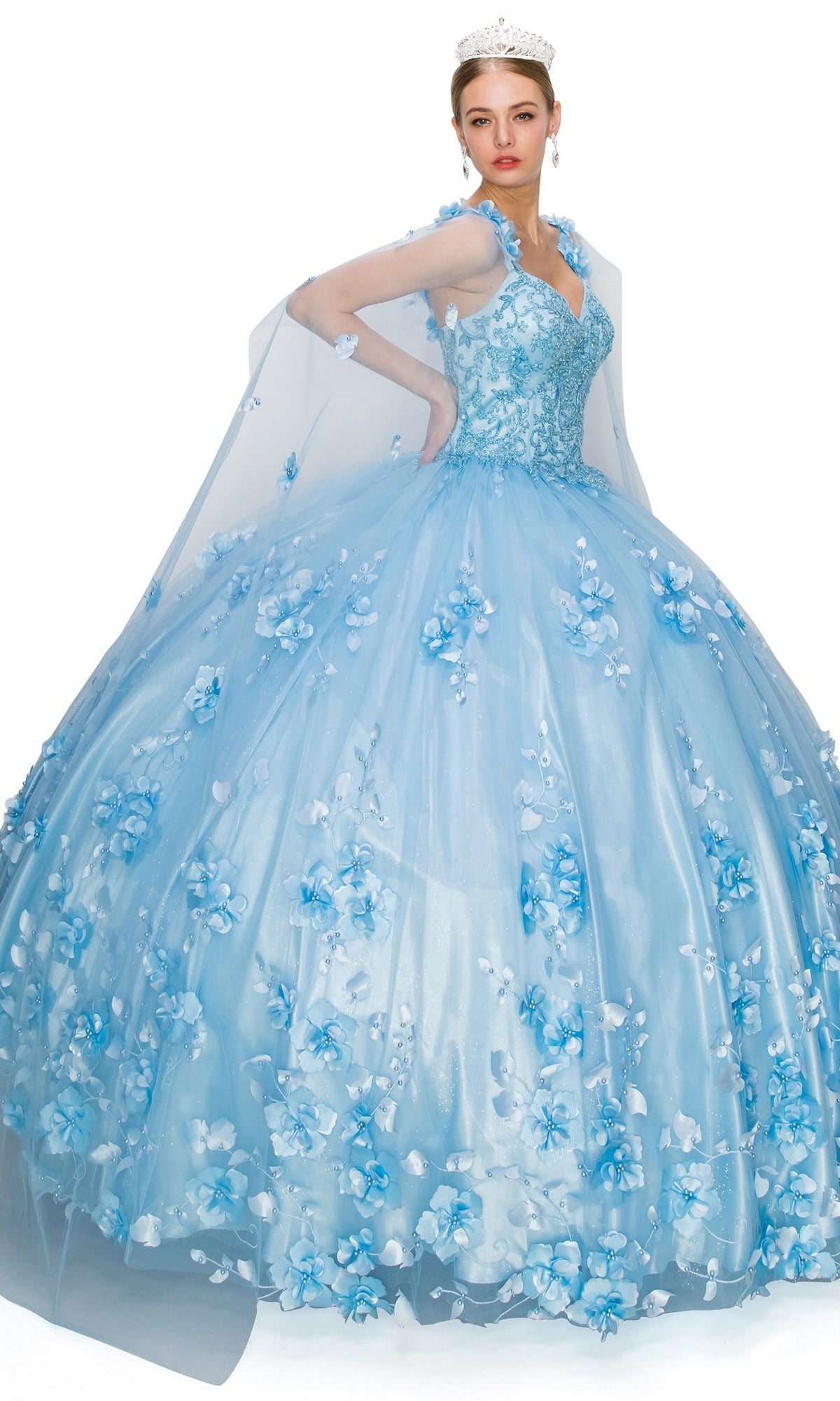 A woman in the Quinceanera Ball Gown 8030J—light blue with 3D floral embellishments, a sheer cape, and a tiara—stands against a white background.