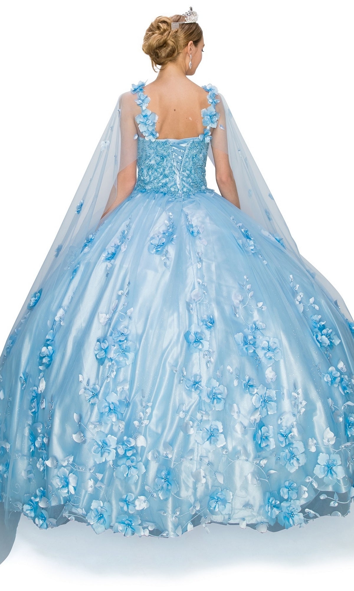 A woman wearing the Quinceanera Ball Gown 8030J, featuring a blue hue, sweetheart neckline, 3D floral details, and a sheer cape, stands with her back to the camera against a white background.