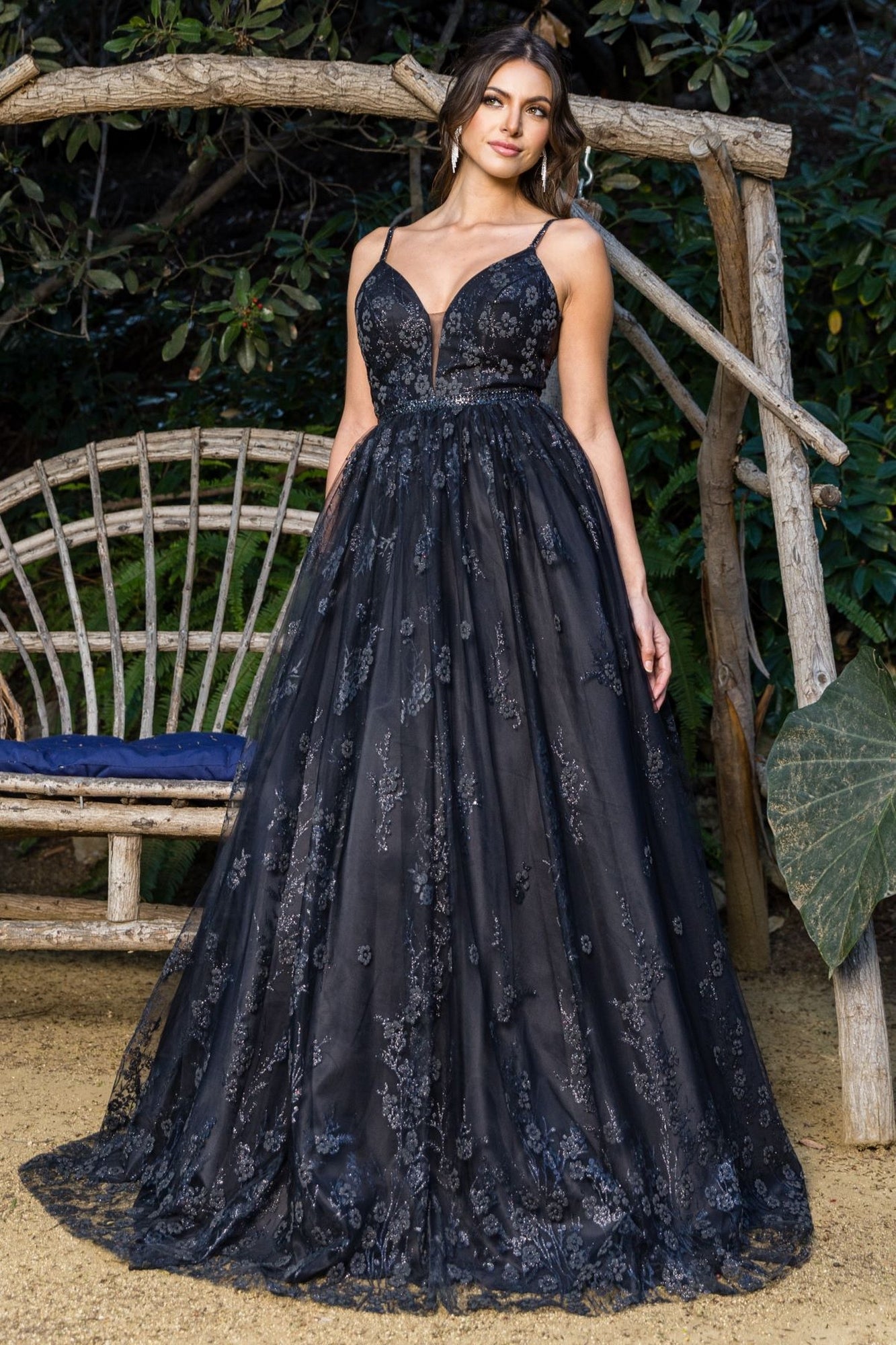 A woman poses outdoors by a rustic wooden bench in the Corset-Back Long Glitter Prom Ball Gown 8039J, a floral embellished dress with a deep neckline, floor-length skirt, and delicate embroidery, surrounded by lush greenery.