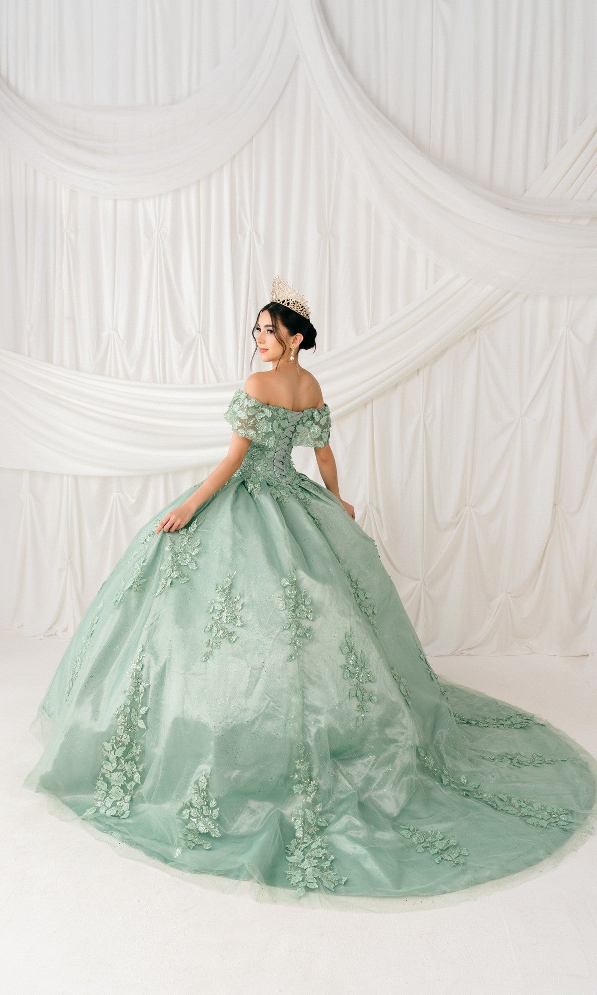 A woman in the Quinceanera Ball Gown 8045J with off-the-shoulder 3D floral detail and a tiara stands facing away from the camera before white draped fabric.