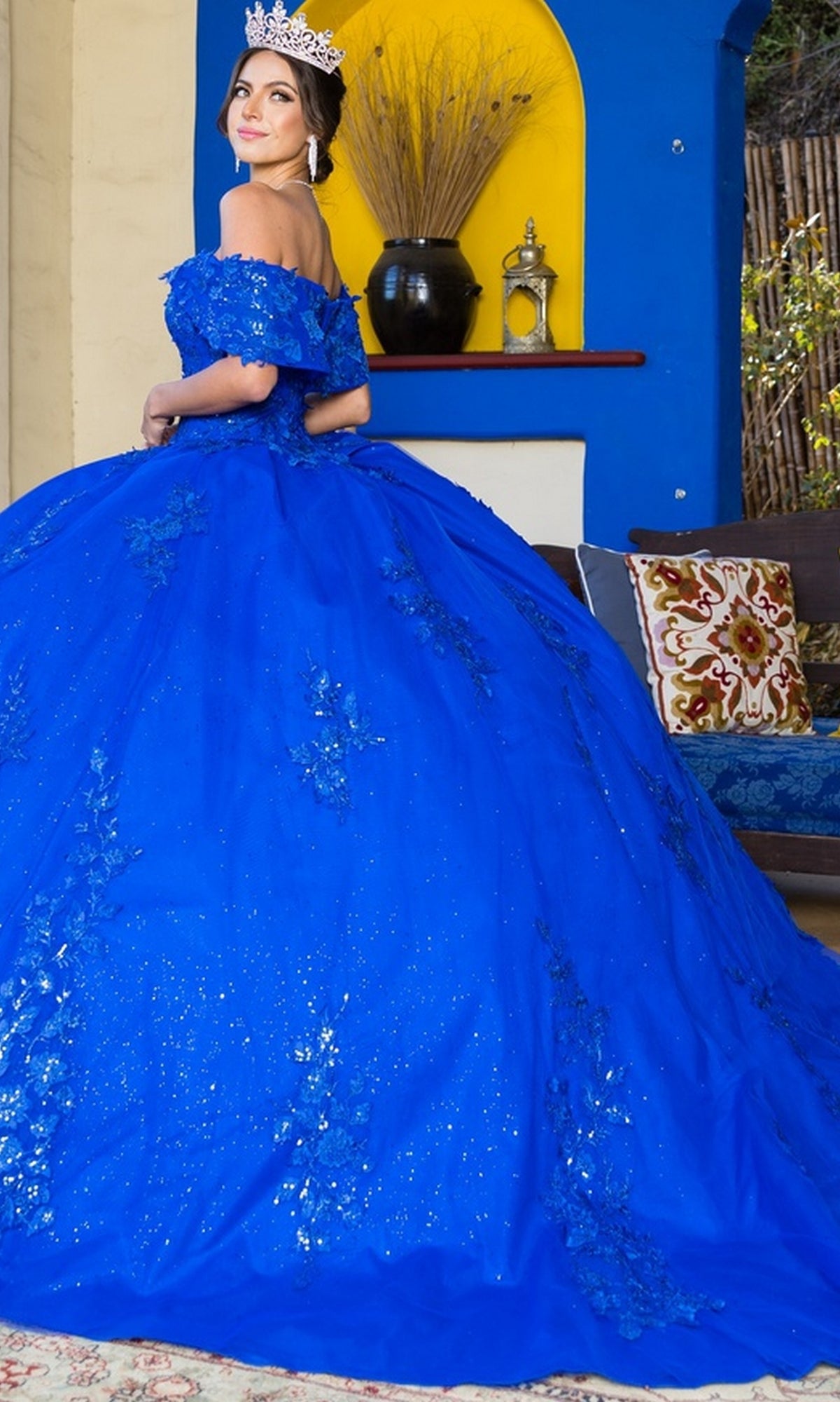 A woman wearing the Quinceanera Ball Gown 8045J, a sparkling royal blue dress with glitter lace embroidery and a tiara, stands indoors looking back over her shoulder.