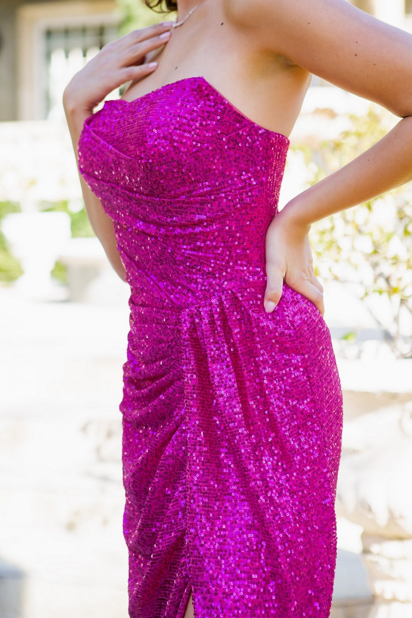 Wearing the Allover-Sequin Long Strapless Prom Dress 8052J in bright pink, a person stands outdoors with one hand on their hip and the other near their neckline.