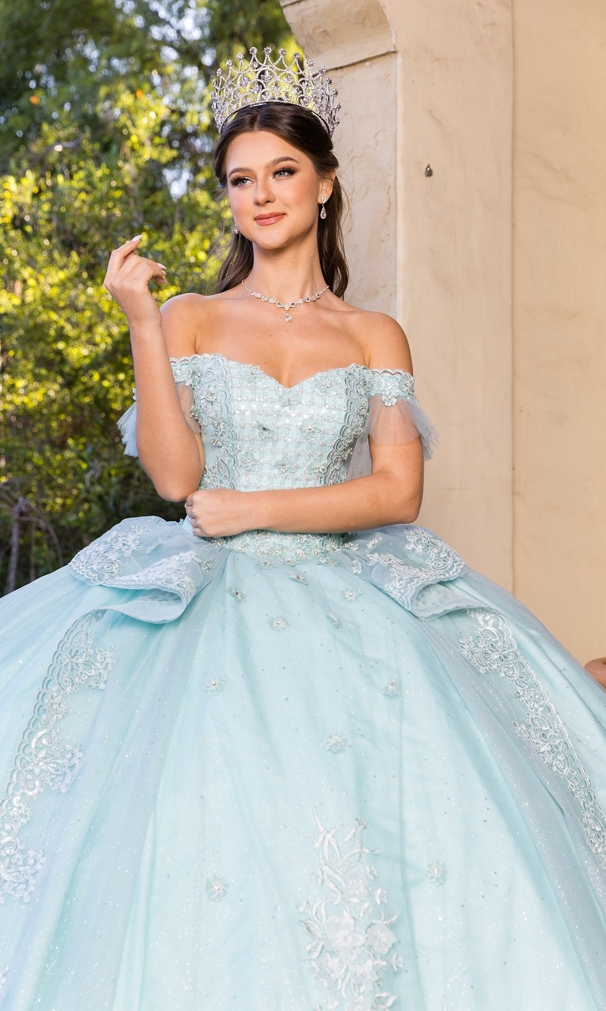 A young woman poses outside near a beige wall in the Quinceanera Dress 8055J by Cinderella Couture, a light blue off-shoulder ball gown with lace embroidery, smiling slightly with one hand raised and wearing a crown.