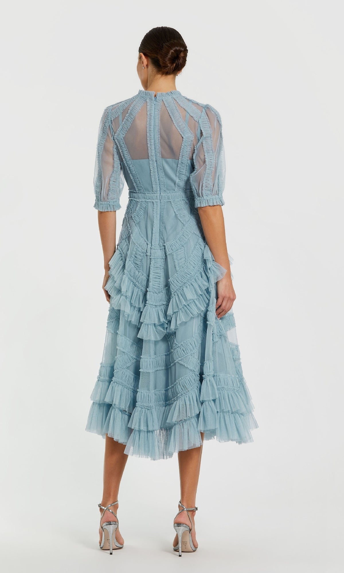 A woman stands with her back to the camera in a Mac Duggal 8058 Ruffled Midi Wedding-Guest Dress, light blue with sheer panels and ruffled layers, paired with heeled sandals; her hair is styled in a neat bun.