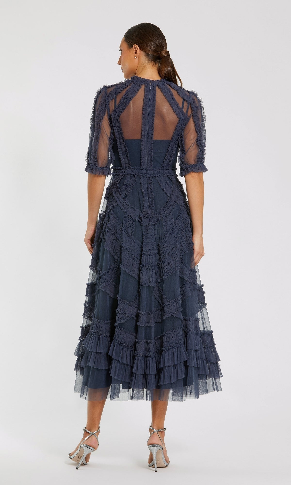 A woman stands facing away, wearing the Mac Duggal 8058 Ruffled Midi Wedding-Guest Dress in navy blue with sheer panels and short sleeves, paired with silver high-heeled sandals.