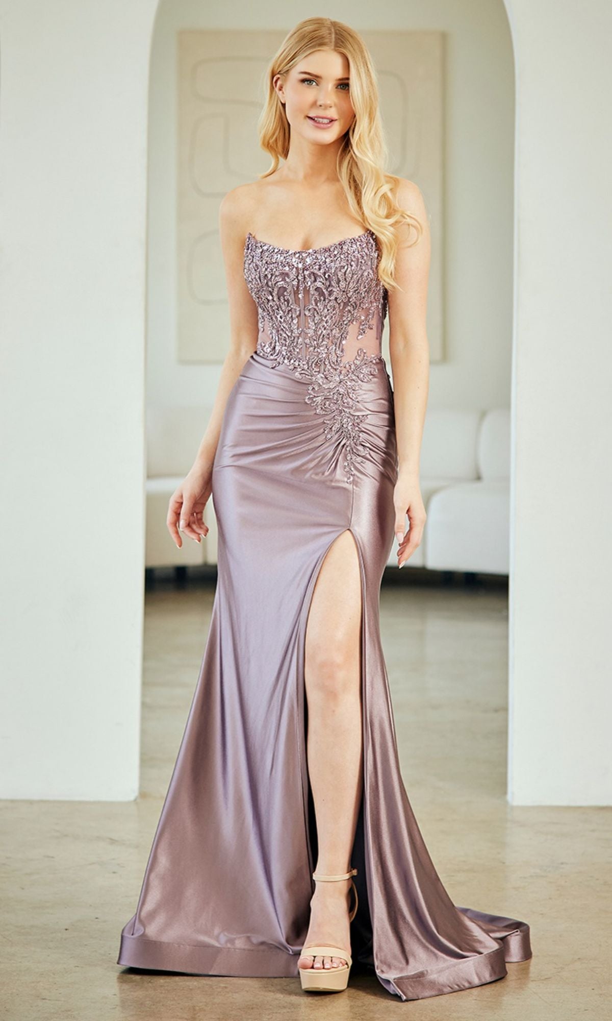 A woman stands indoors on a polished floor wearing the Sheer-Corset Long Prom Gown: Amelia Couture 8059, a lavender satin strapless dress with an embellished corset bodice, high slit, and lace details.