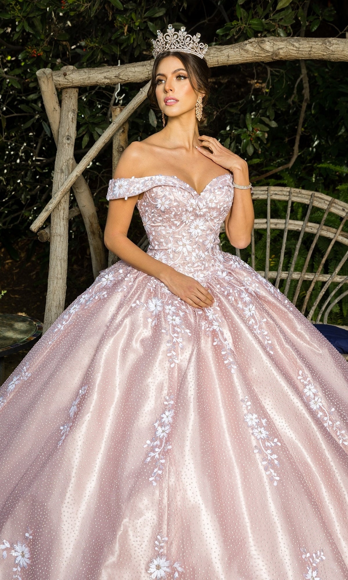A woman wearing a silver tiara and the Quinceanera Ball Gown 8060J—an off-the-shoulder dress with pink embossed tulle and floral embroidery—stands before wooden branches and foliage.