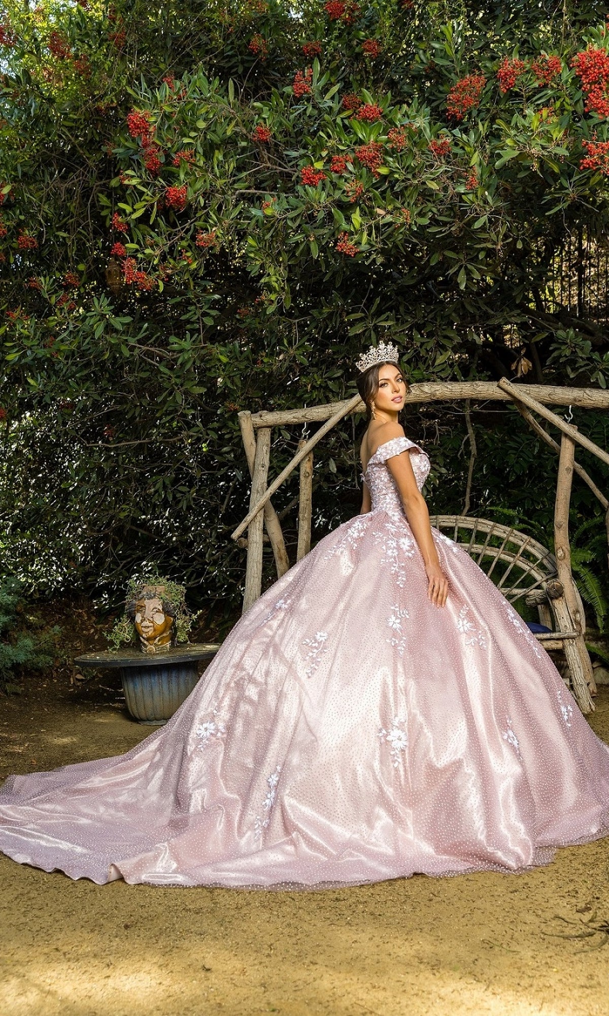 A young woman wearing the Quinceanera Ball Gown 8060J, a pink off-the-shoulder dress, and a crown stands in front of a rustic wooden swing surrounded by greenery and red flowers.