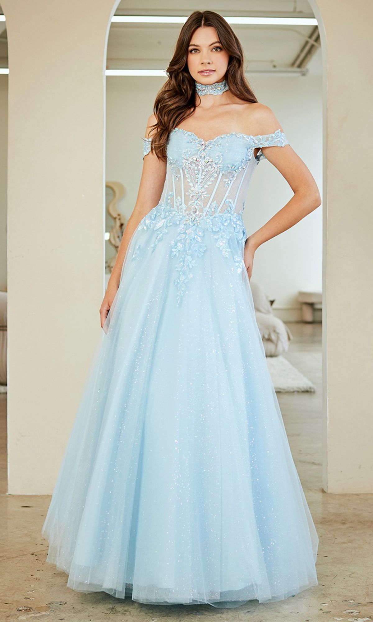 Wearing the Amelia Couture 8064 long prom dress in light blue with an off-the-shoulder design, sheer corset bodice, floral appliqué, and matching choker, a woman stands in a softly lit room.