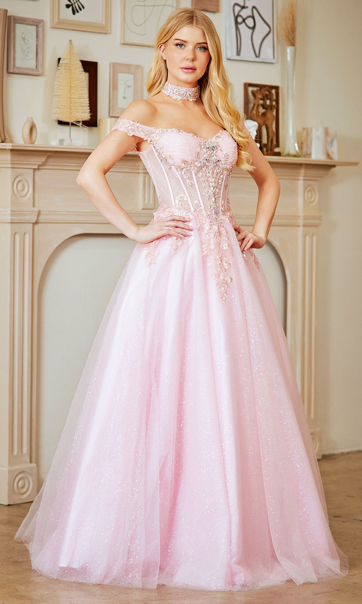 A woman in the Amelia Couture 8064 long prom dress, light pink and off-the-shoulder with a fitted embellished bodice, stands indoors before a cream-colored fireplace.