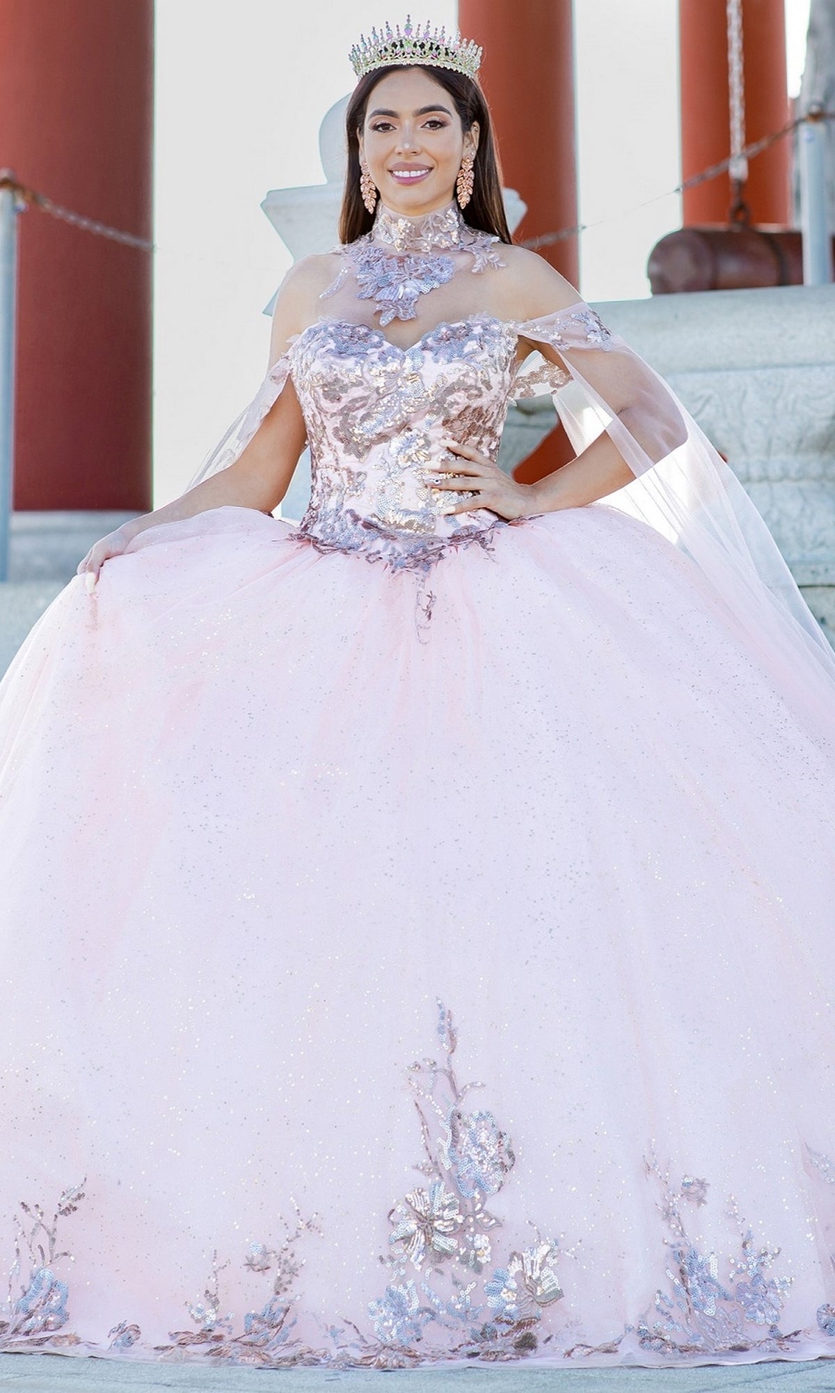 A young woman poses outdoors in front of stone steps wearing the Quinceanera Ball Gown 8064J, featuring sparkling pink lace embroidery with floral details, an off-the-shoulder design, and detachable sleeves, accessorized with a tiara.