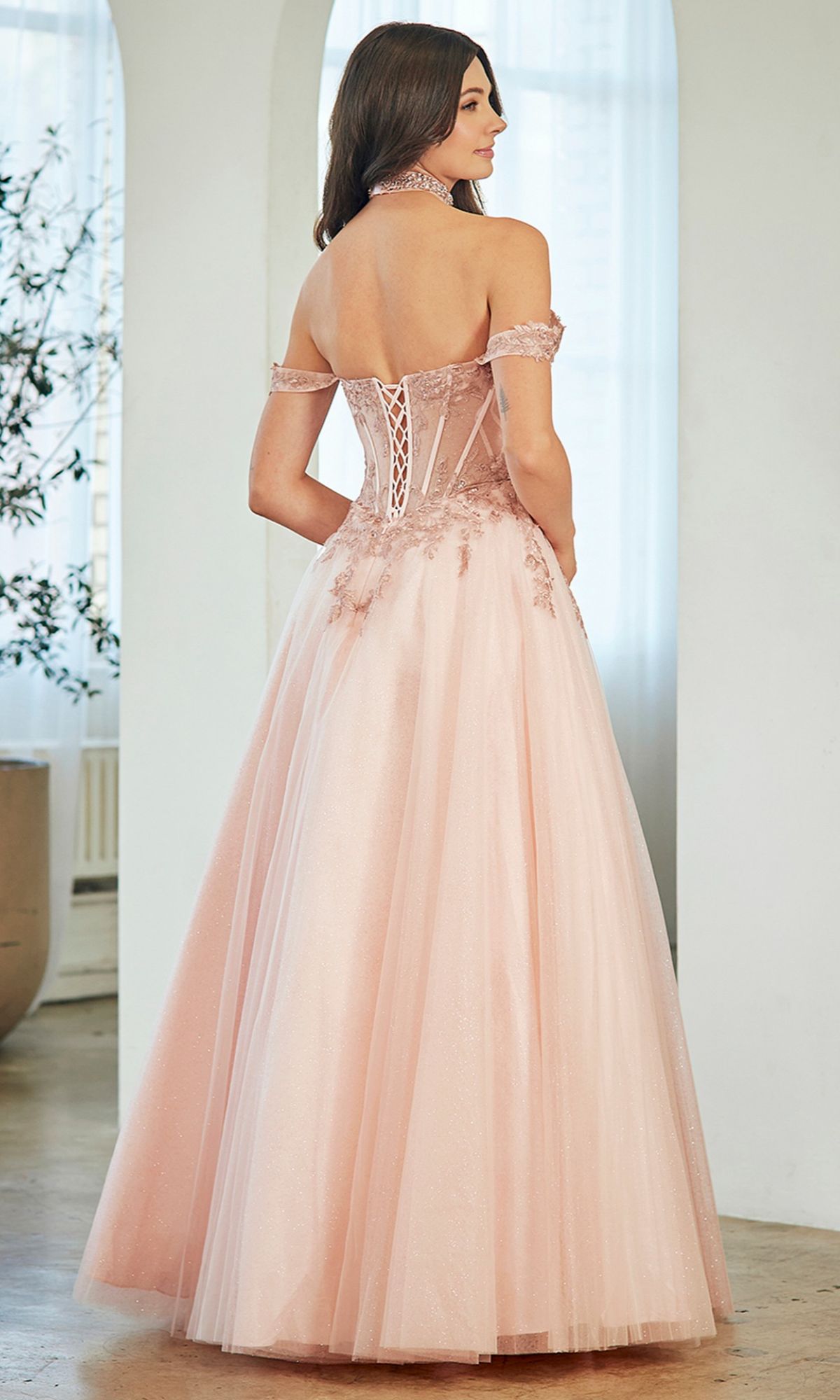 Woman wears the Amelia Couture 8065 long prom dress in pink, featuring an off-the-shoulder design, lace detailing, corset back, and glitter tulle. She stands indoors facing away from the camera.