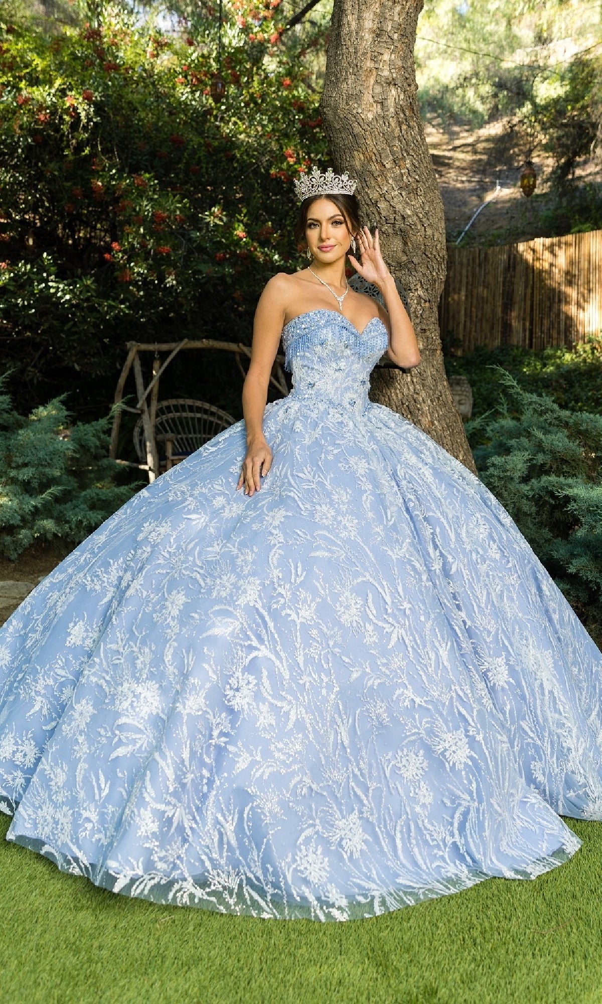 A woman in the Cinderella Couture 8071J quinceanera dress—a light blue, strapless ball gown with white embroidery and beaded fringe—wears a tiara and stands outdoors beside a tree.