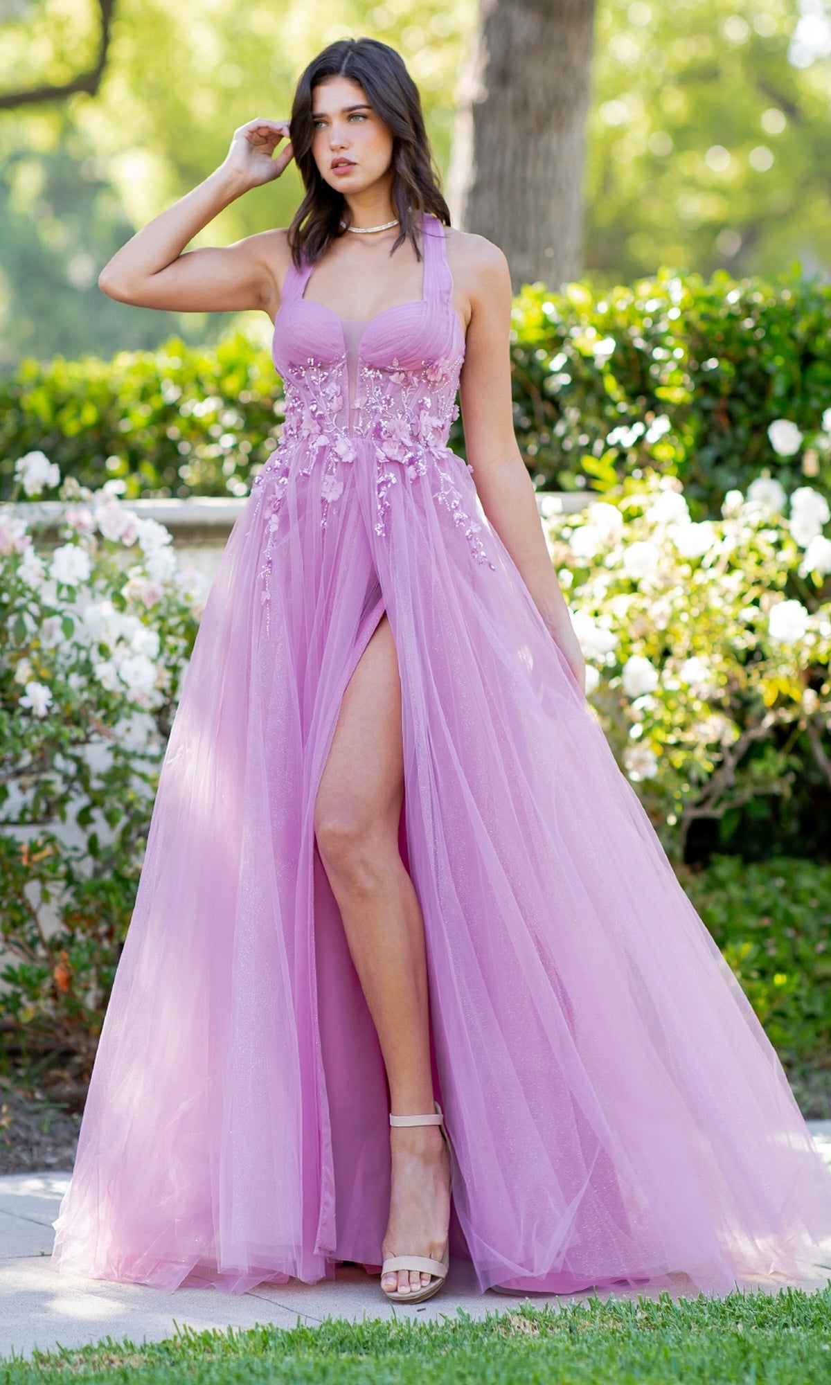 A woman stands outdoors in greenery and white flowers, wearing the Cinderella Couture 8076J—a sleeveless light purple long halter prom dress with floral embellishments and a high slit.