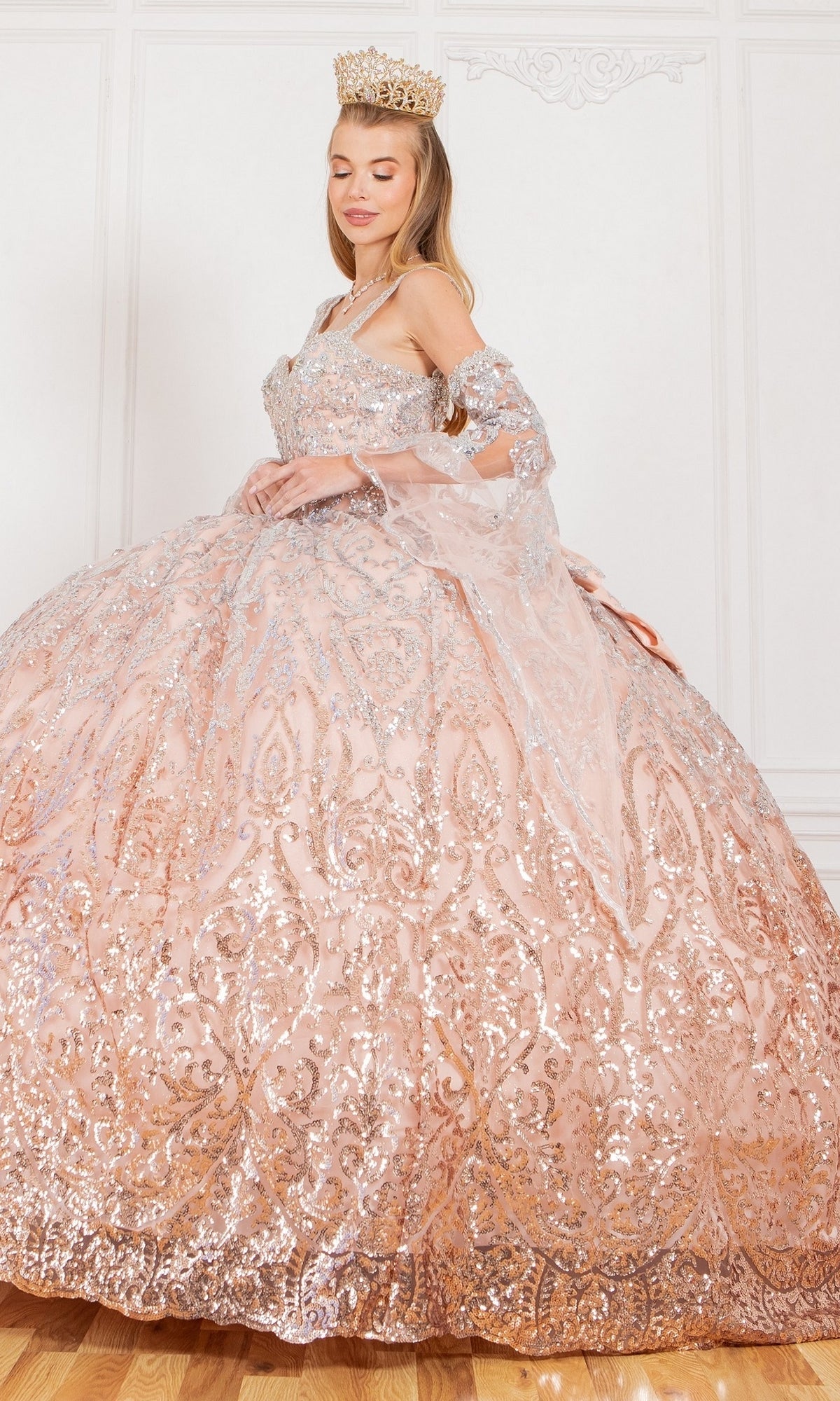 A woman in the Quinceanera Ball Gown 8080J, adorned with large crystal beads, silver sequins, and a crown, stands before a white wall.