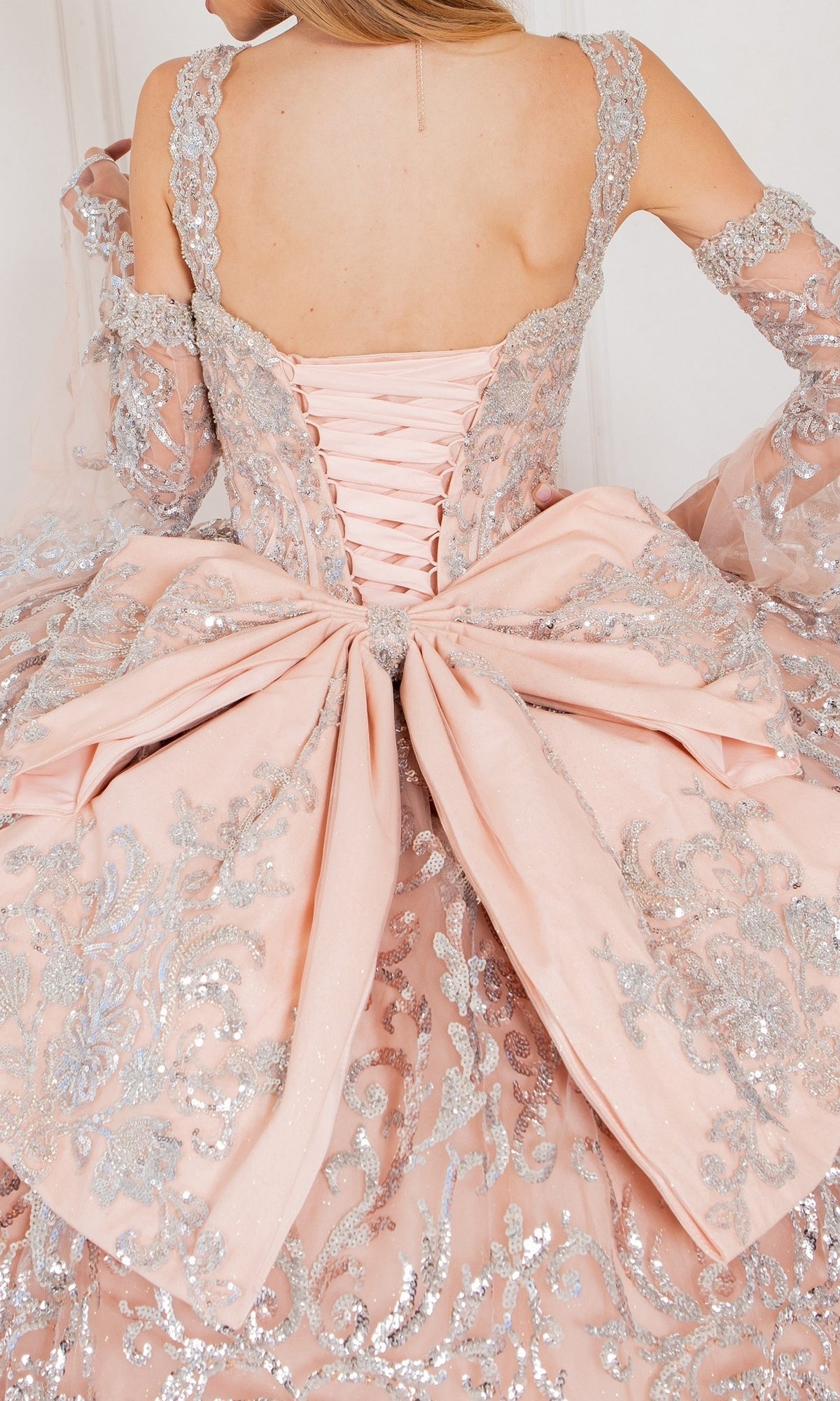 A close-up of a person wearing the Quinceanera Ball Gown 8080J, featuring a pink ombre design, silver embroidery, lace-up back, and a large decorative bow at the waist.