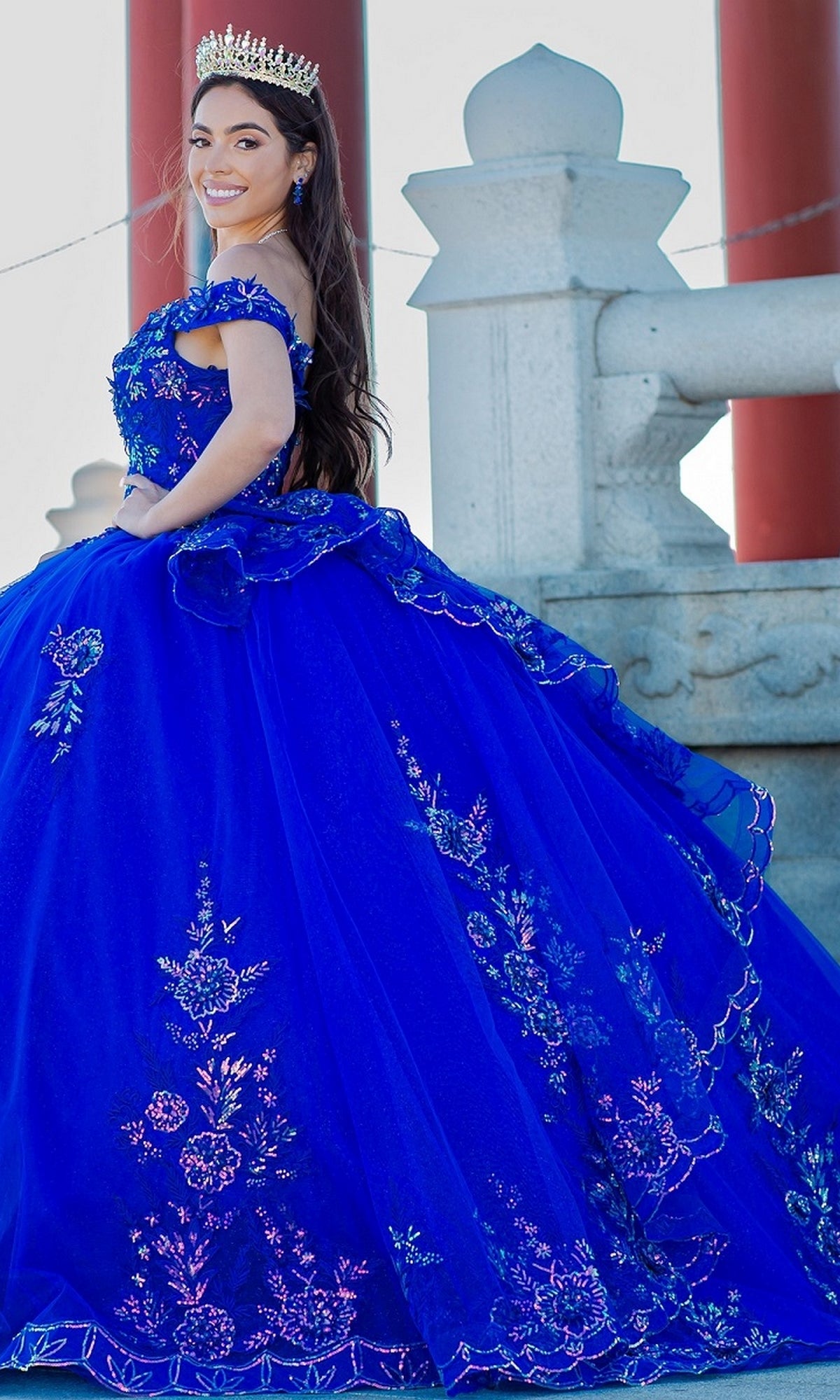 A woman in the Quinceanera Ball Gown 8100J, featuring an off-the-shoulder design and iridescent beaded details, smiles and poses outdoors beside a stone railing while wearing a crown.