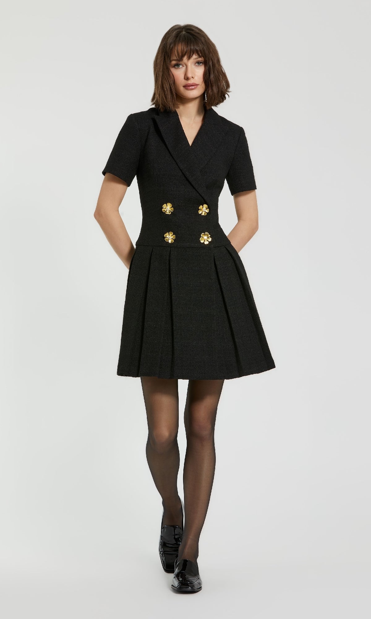 A woman models the Mac Duggal 81038 Short Tweed Blazer Dress with gold buttons, paired with sheer black tights and black shoes, against a plain white background.