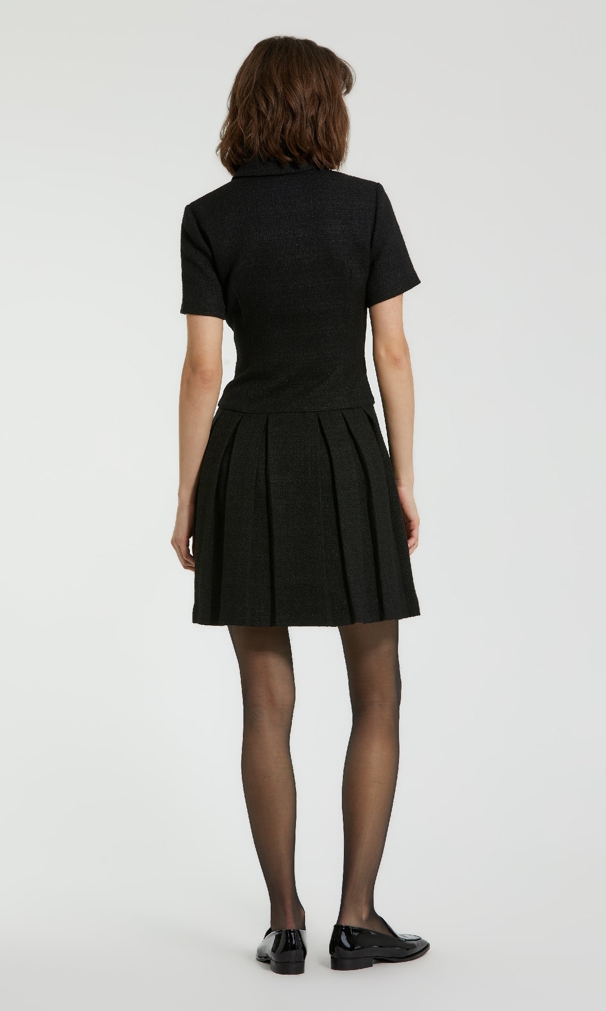 A person stands facing away, wearing the Mac Duggal 81038 Short Tweed Blazer Dress with a pleated A-line skirt, black sheer tights, and black shoes against a plain background.
