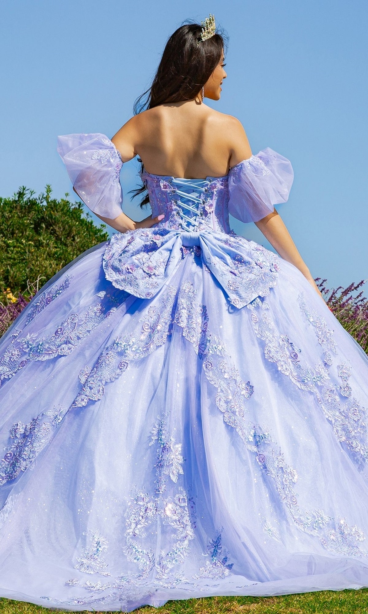 A woman stands outdoors facing away, wearing the Quinceanera Ball Gown 8115J in light purple with floral embellishments, removable puff sleeves, and a lace-up back.