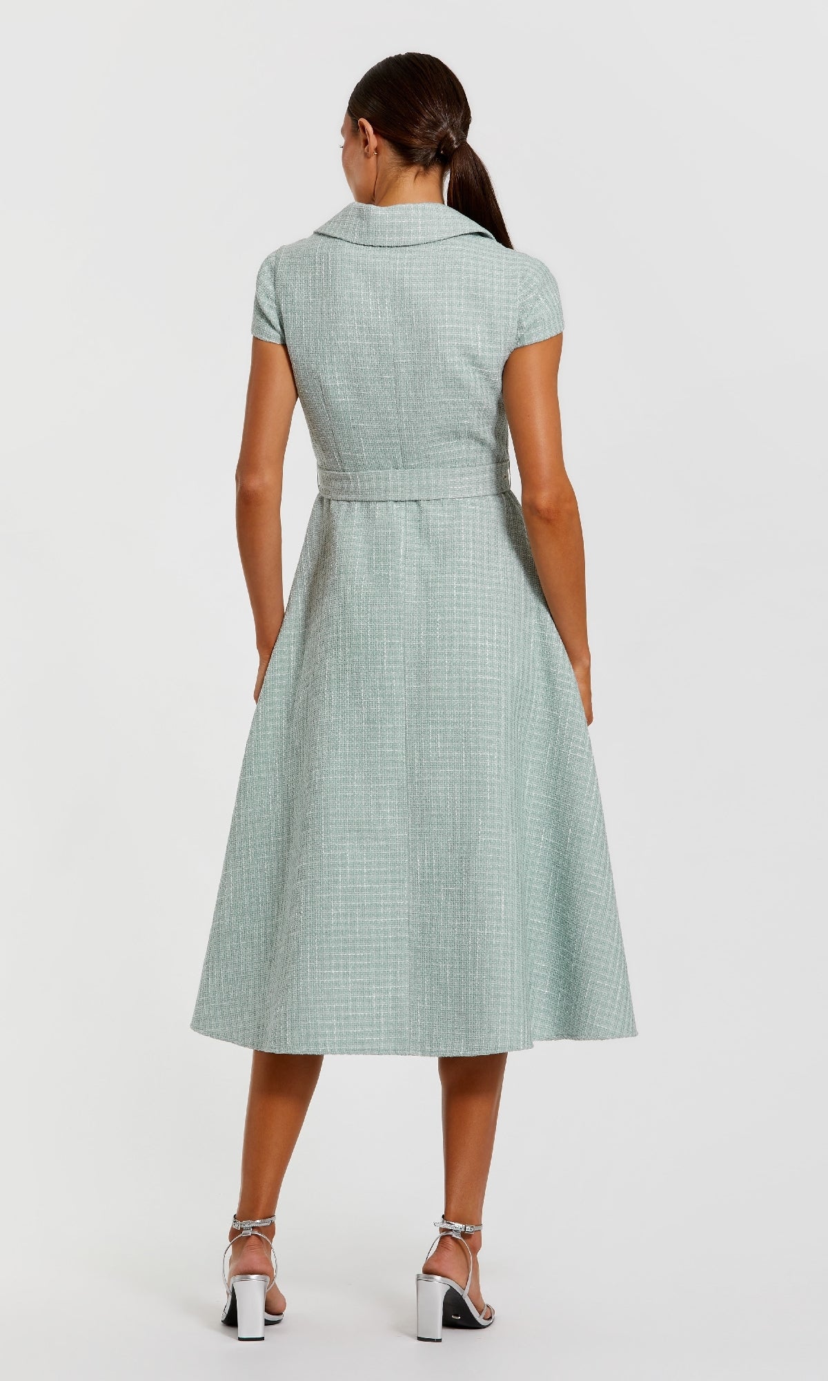 A woman faces away from the camera in a Collared Midi Green Career Dress: Mac Duggal 81199 with short sleeves and a belt, styled with white heeled sandals against a plain white background.