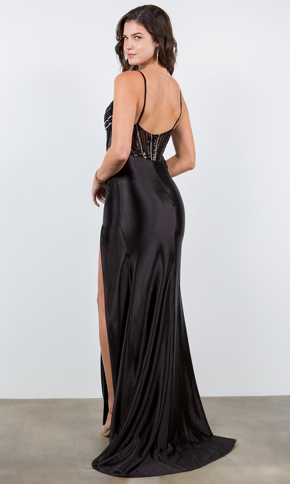 A woman in the Sequin-Bodice Prom Dress: Cinderella Couture 8122J, featuring a sleeveless, floor-length design with a v-neckline, high slit, and open back, stands on a plain background, looking over her shoulder.