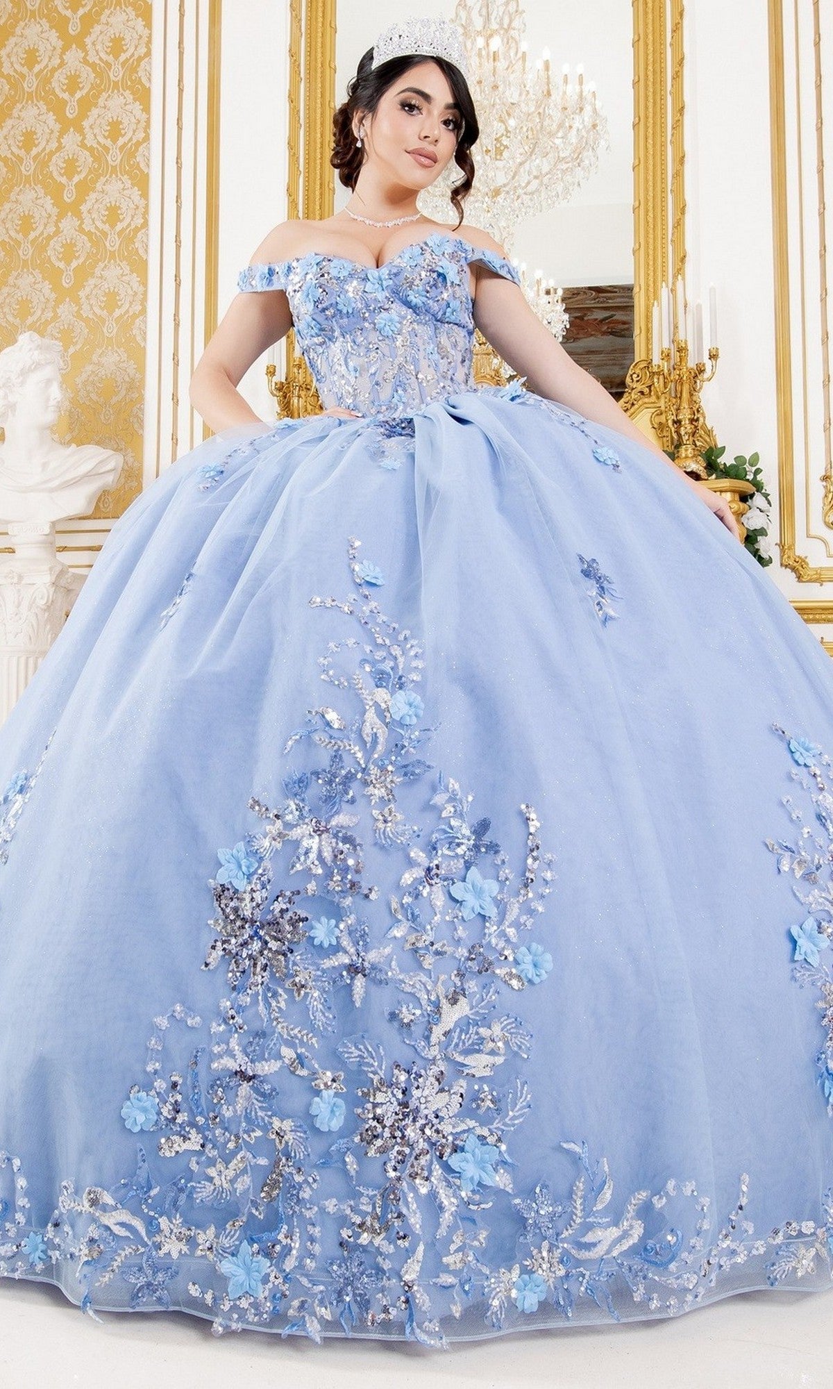 A young woman wears the Quinceanera Ball Gown 8125J, featuring an off-the-shoulder design, corset lace-up bodice, and colorful embellishments, while posing indoors with a tiara before a gold-framed mirror.