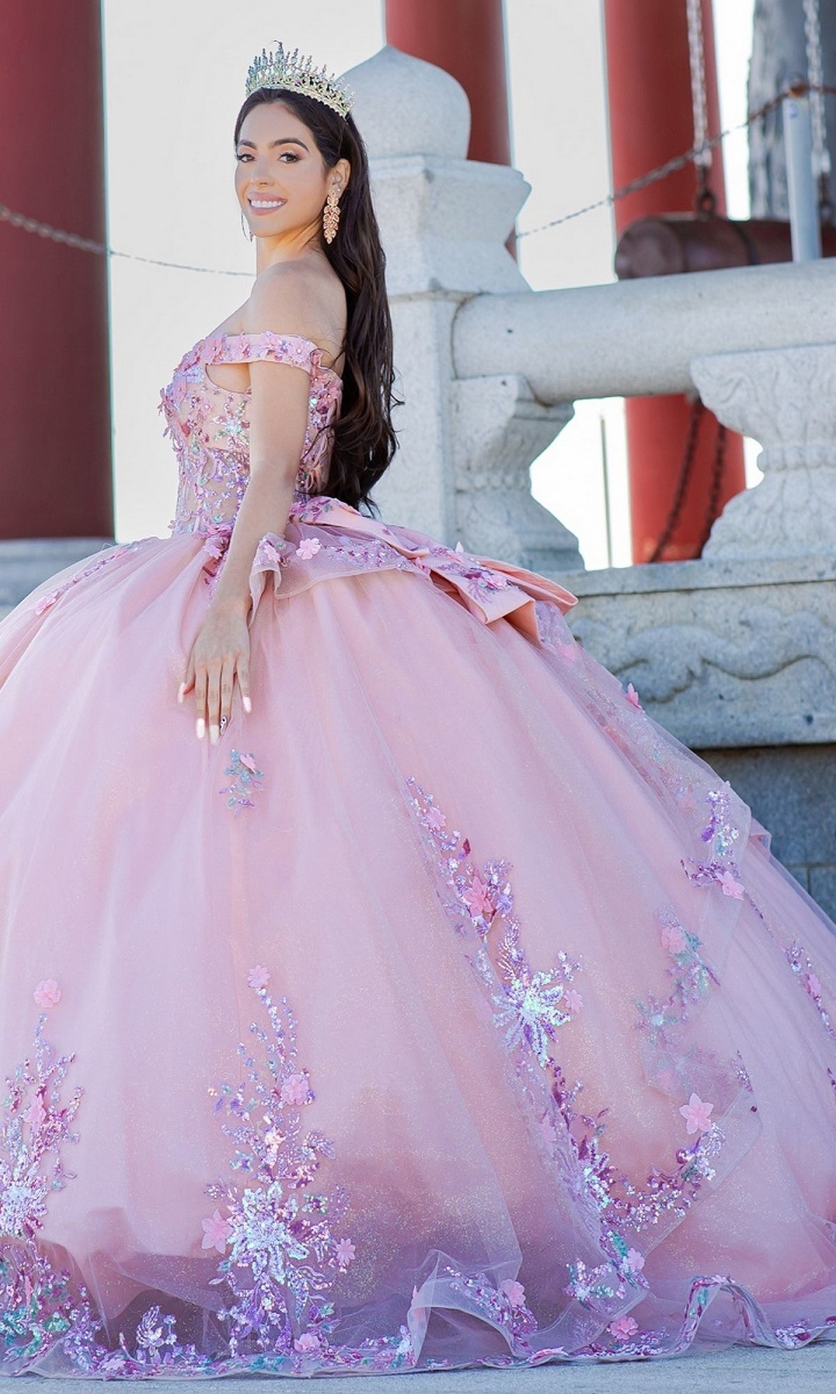 A young woman in the Quinceanera Ball Gown 8125J—an off-the-shoulder, pink floral dress—wears a tiara and poses outdoors before a stone railing and large red columns.