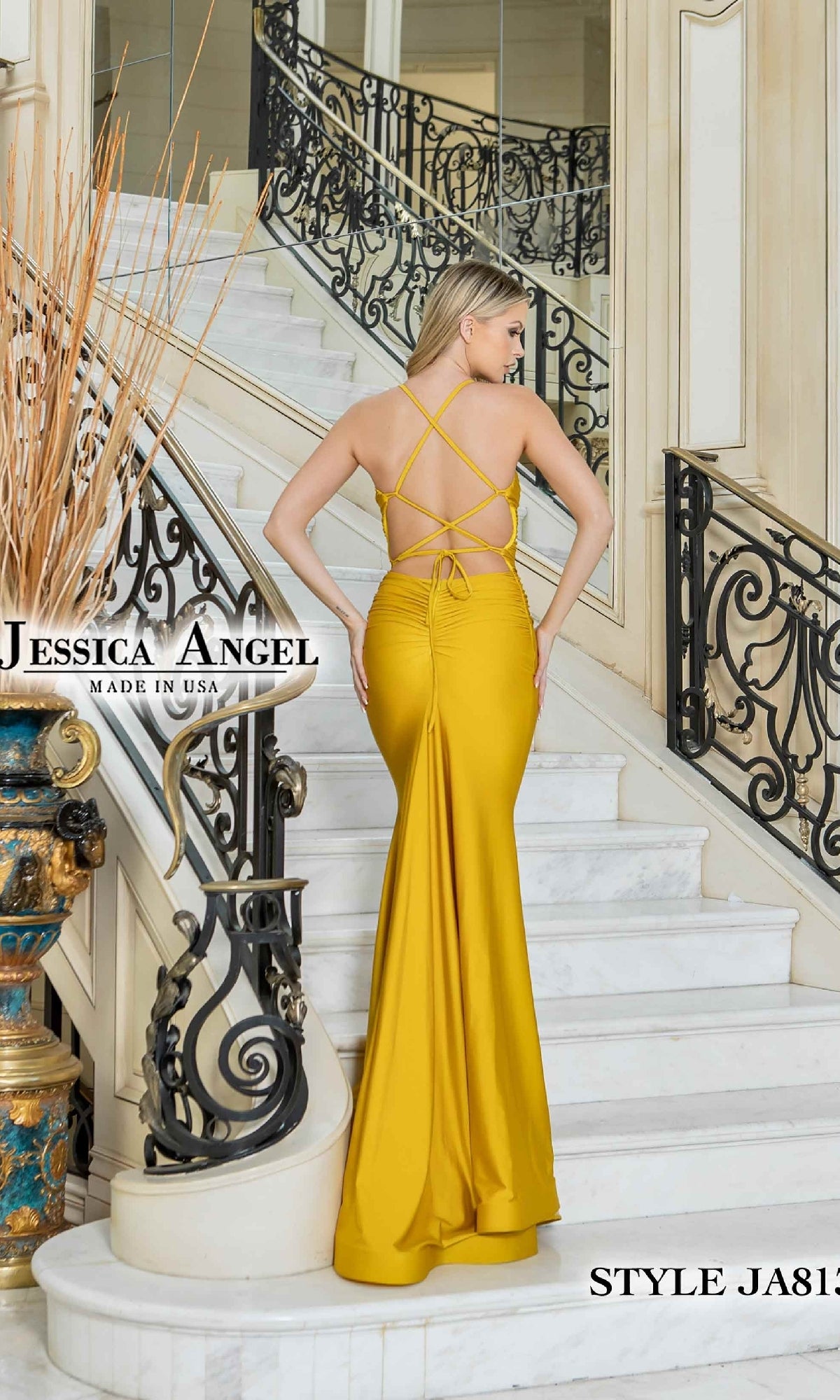 A woman in a yellow Lace-Up Classic Long Prom Dress: Jessica Angel 813 stands on a white marble staircase with black railings. The fitted gown features crisscross straps, a backless design, and a classic silhouette.
