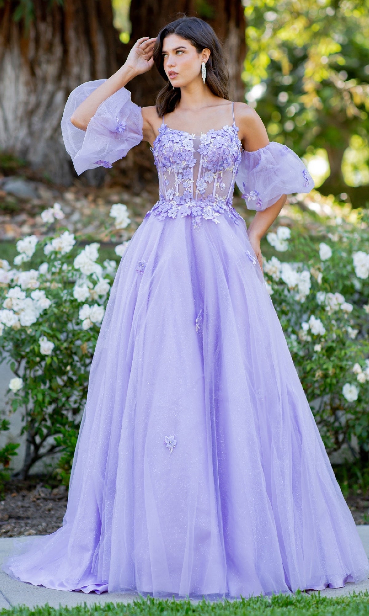 A woman stands outdoors among white flowers and greenery in the Purple Prom Ball Gown: Cinderella Couture 8130J, featuring a long purple silhouette with sheer puff sleeves and floral appliqué details.