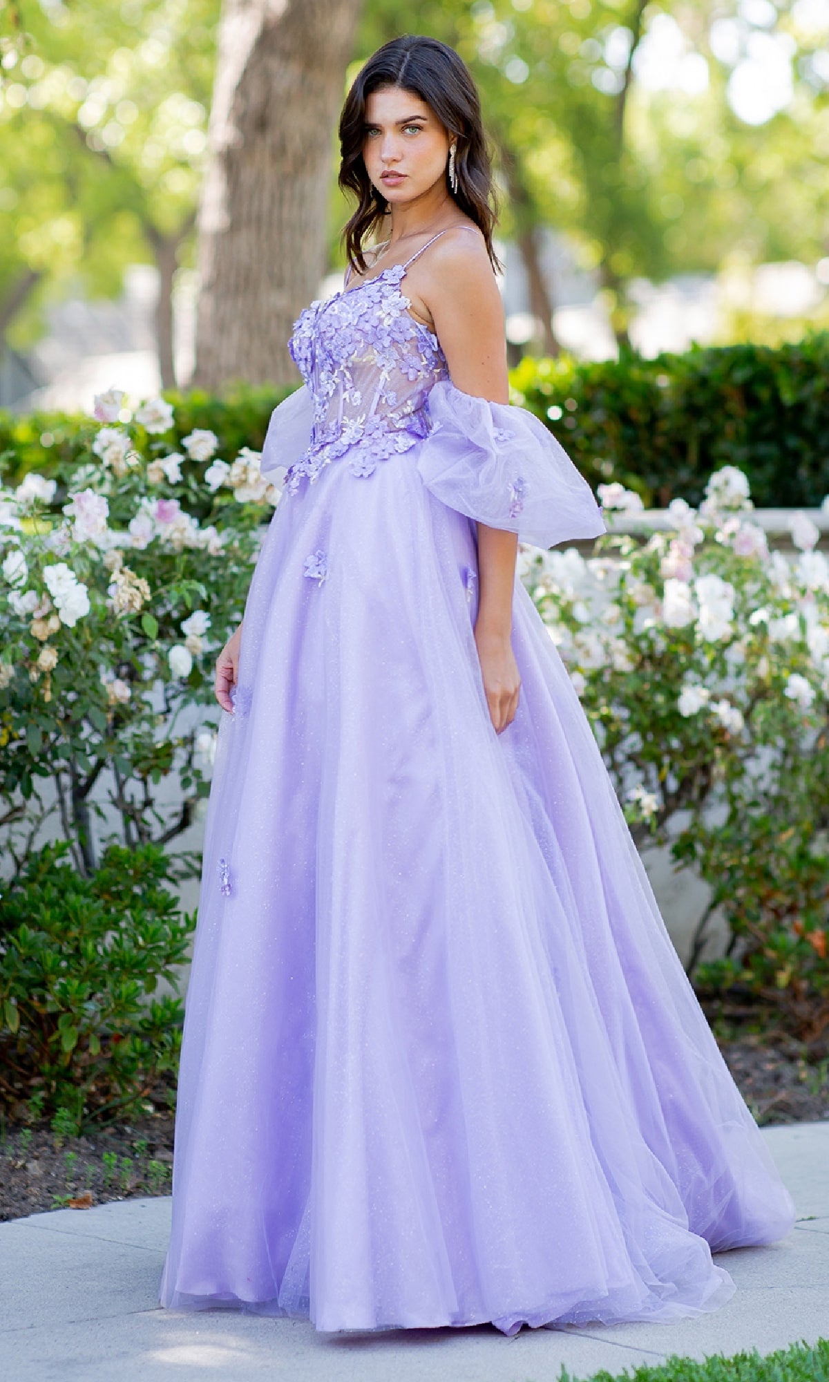 A woman stands outdoors in the Cinderella Couture 8130J Purple Prom Ball Gown, featuring floral appliqué and a sheer bodice, surrounded by greenery and white flowers.