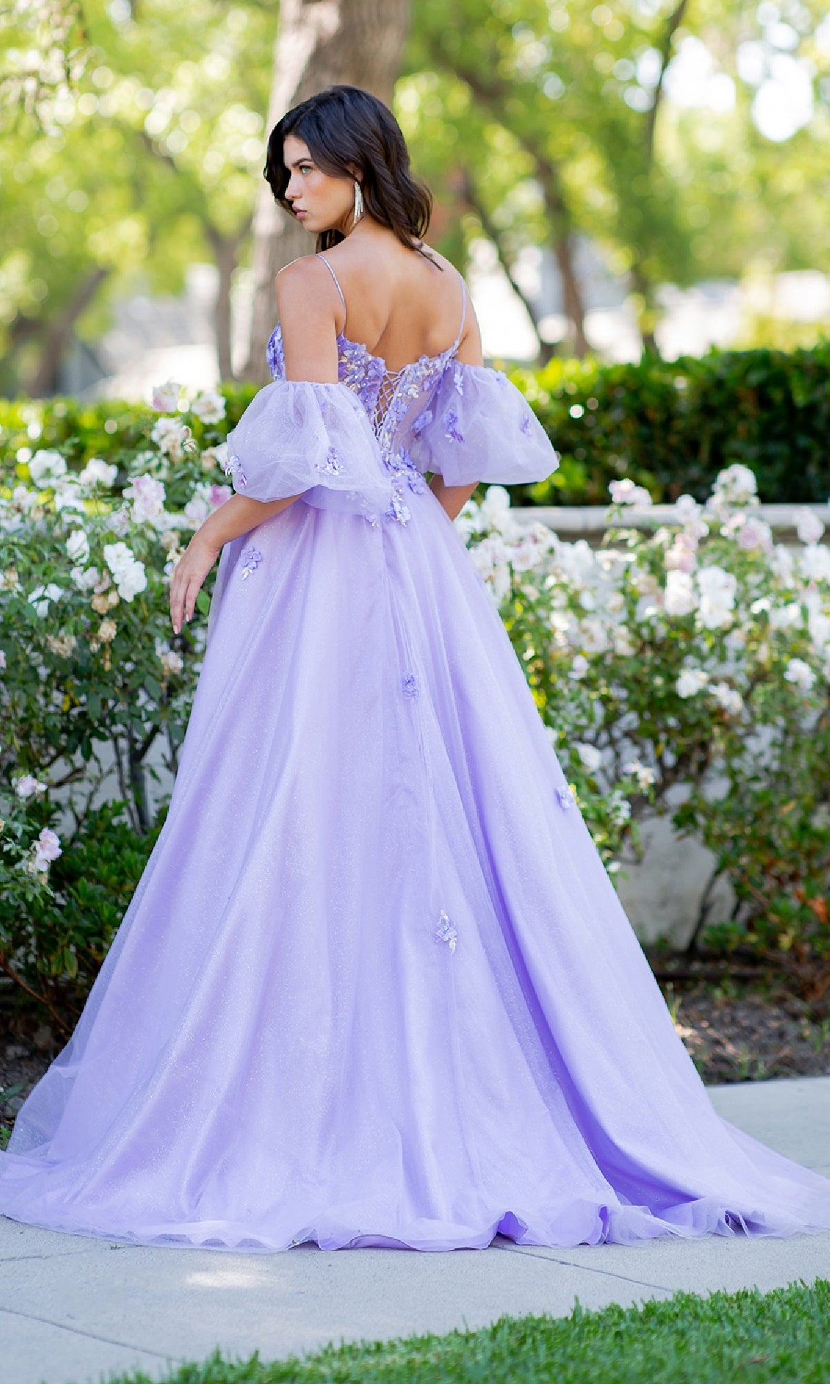 A woman stands outdoors among bushes and flowers, wearing the Cinderella Couture 8130J Purple Prom Ball Gown featuring a lace-up back and puffed sleeves.