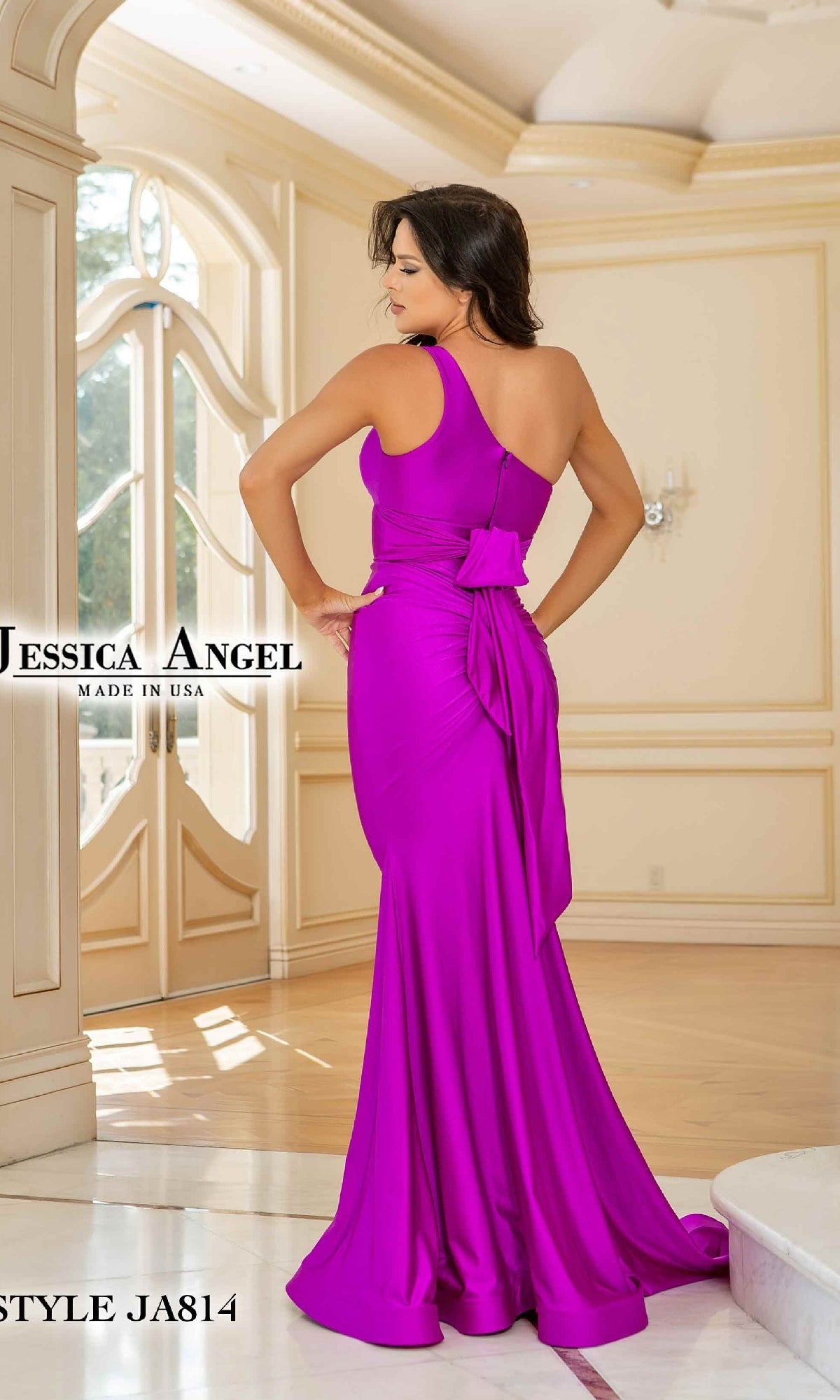 A woman stands indoors near large windows, wearing the Jessica Angel 814—a bright purple, one-shoulder prom gown featuring a long silhouette and a stylish bow at the back.