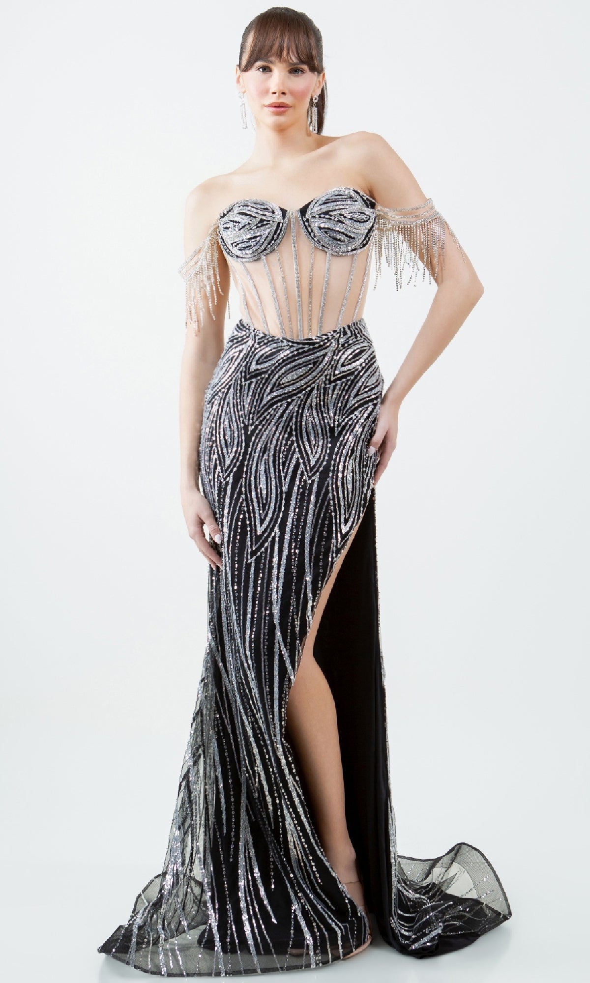 A woman models the Cinderella Couture 8147J—an off-the-shoulder, floor-length black prom dress with silver sequin patterns, a sheer corset bodice, high slit, and beaded details—standing against a plain background.