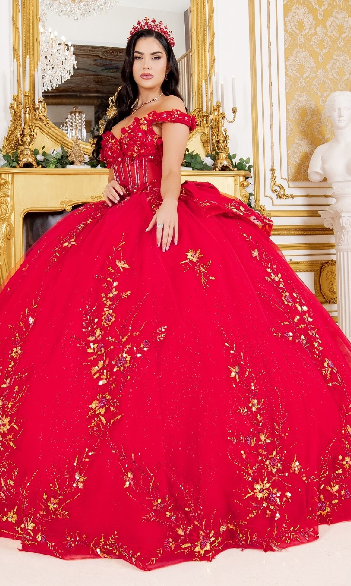 A woman stands in an ornate room wearing the Cinderella Couture 8160J off-the-shoulder red quinceanera ball gown with gold floral embroidery and a matching headpiece.