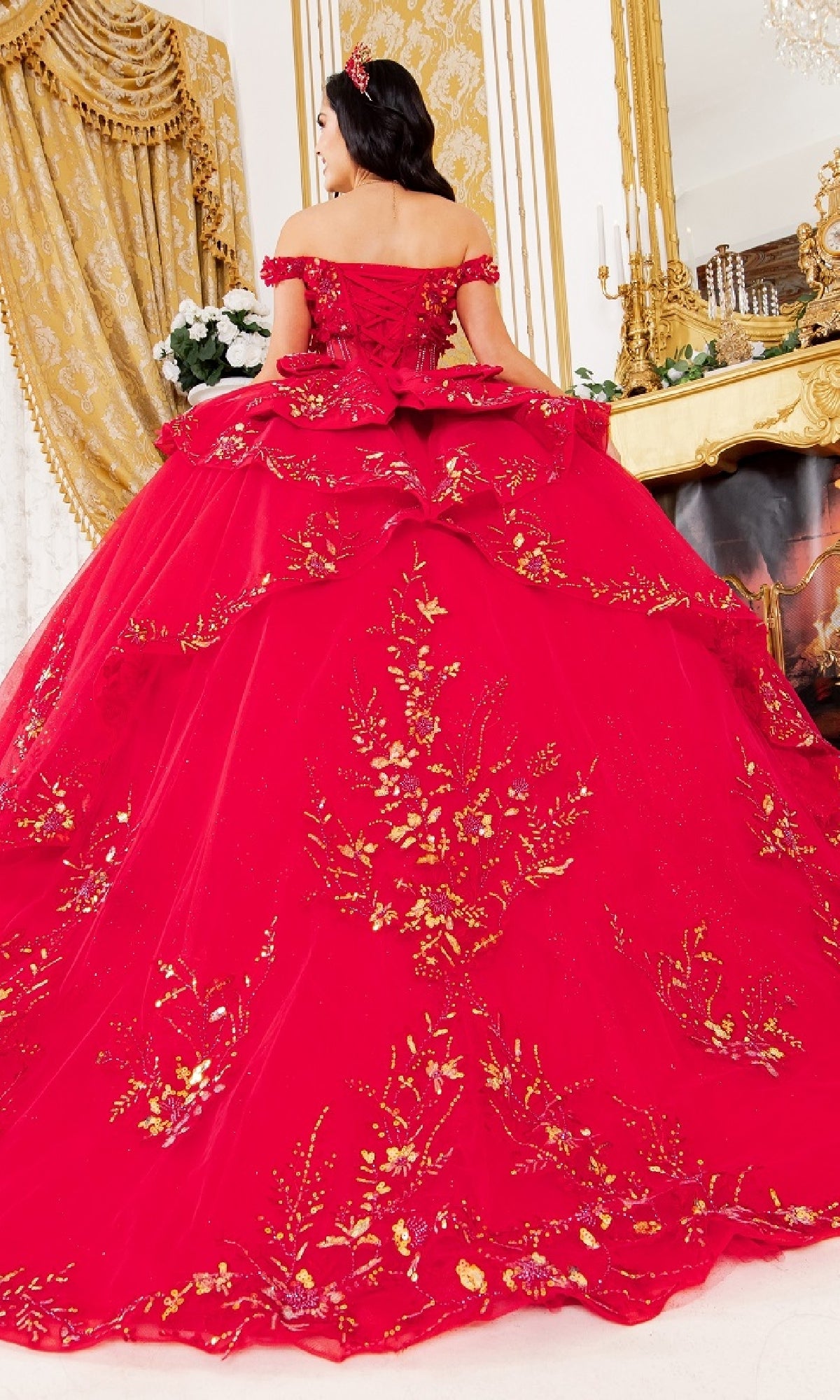A woman faces away from the camera in an elegant gold-decorated room, wearing the Cinderella Couture 8160J quinceanera dress—an ornate red ball gown adorned with gold embroidered lace appliques.