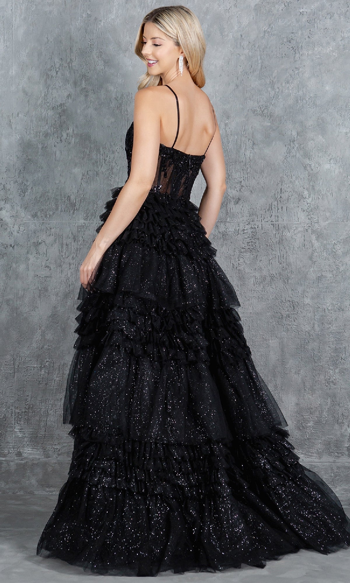 Woman in a sleeveless, floor-length Cinderella Couture 8163J black glitter prom ball gown with ruffled skirt and beaded corset bodice, standing sideways before a gray textured background.