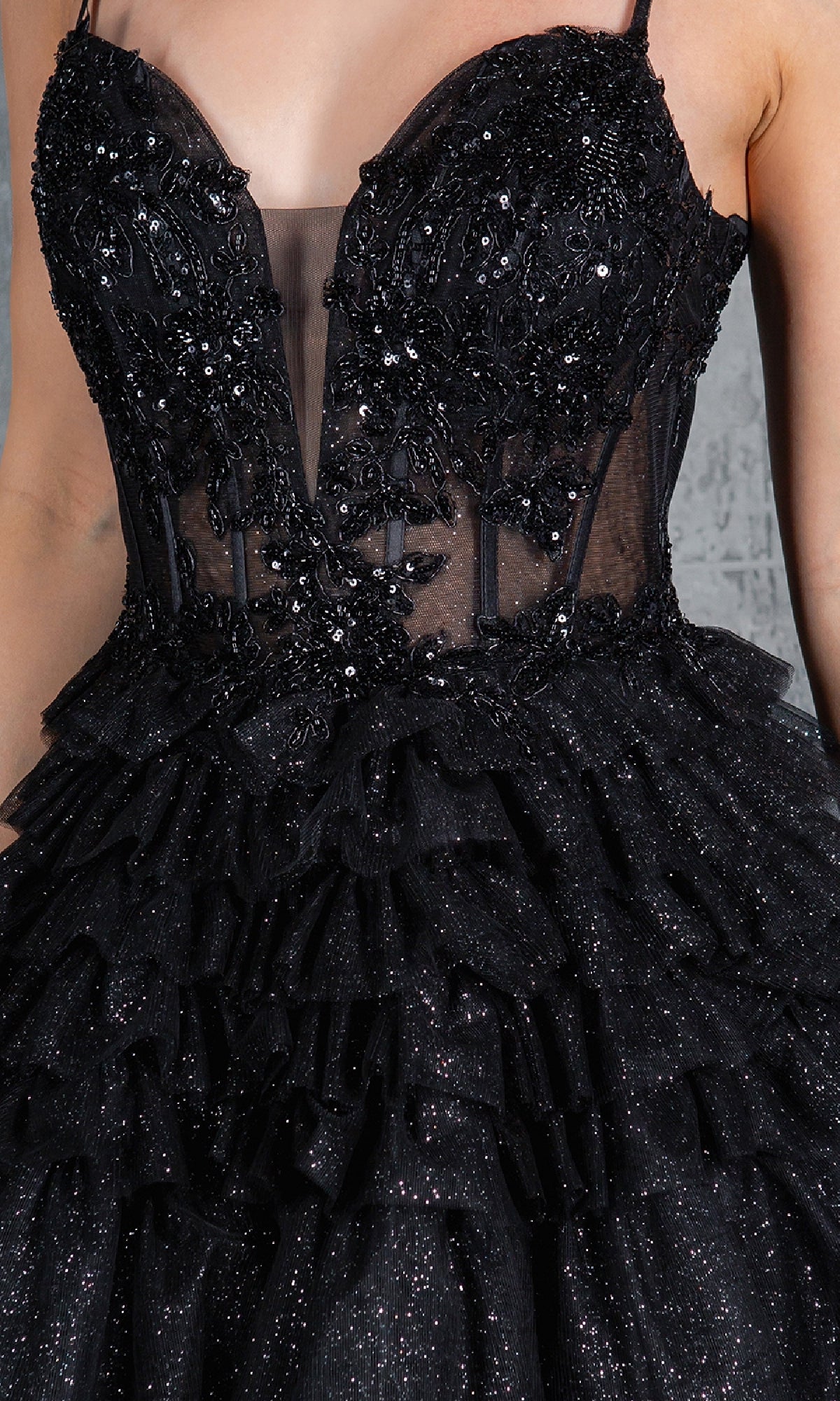A close-up of a person wearing the Glitter Prom Ball Gown: Cinderella Couture 8163J—a sleeveless black dress with sequins, lace appliqué, sheer panel, and tiered ruffled layers, evoking a glamorous A-line silhouette.