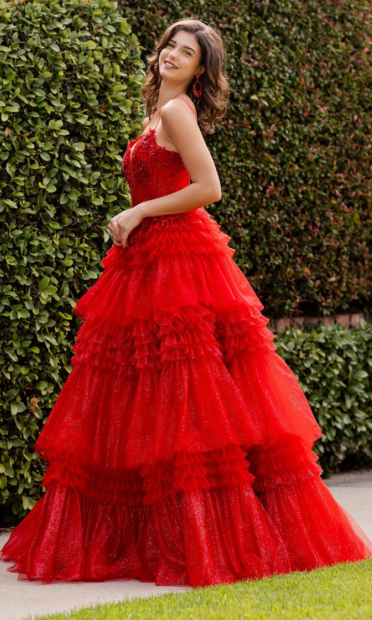 A woman stands outdoors by green hedges, smiling in the Cinderella Couture 8163J Glitter Prom Ball Gown—a sleeveless, tiered red dress with ruffles and sparkling details.