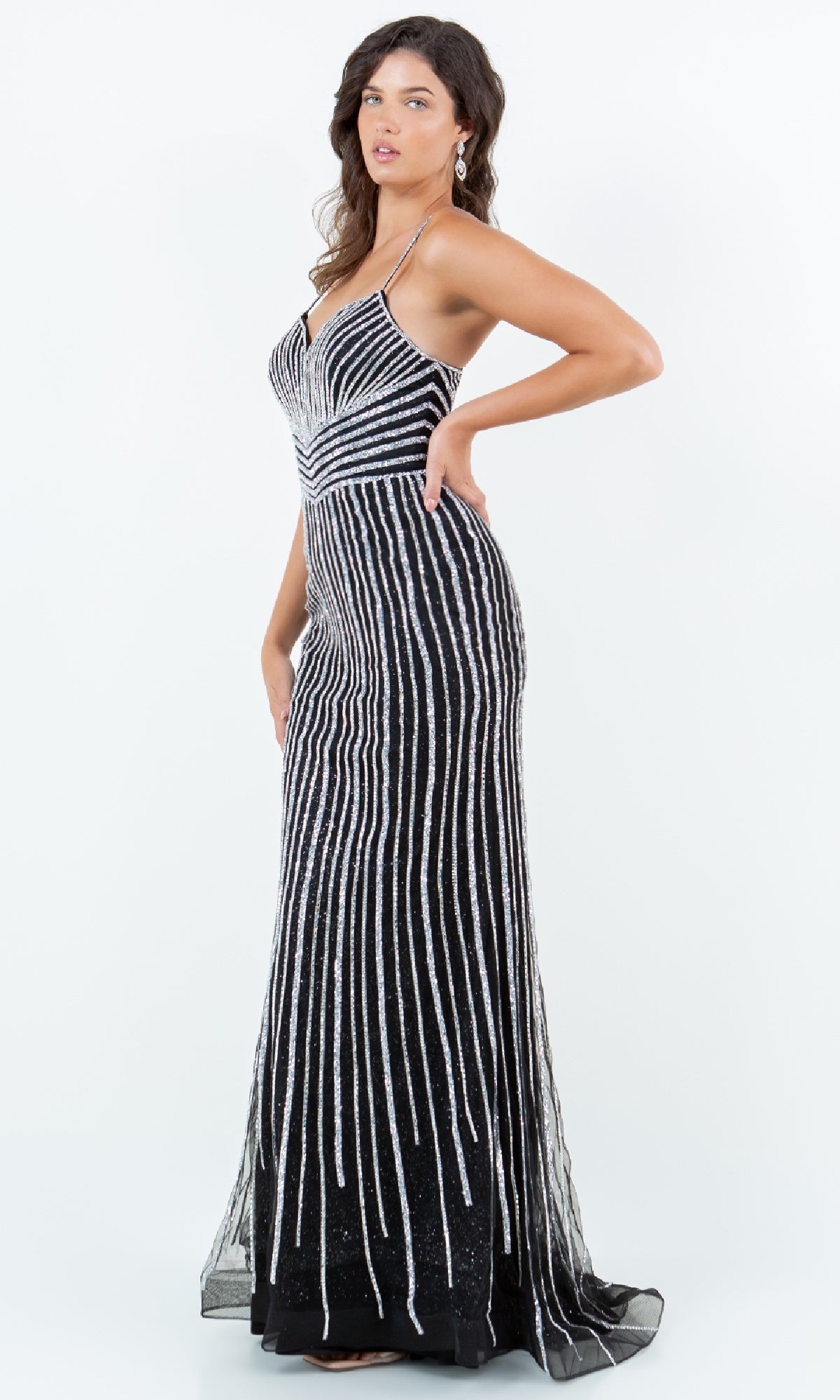 A woman models the Bead-Striped Prom Gown: Cinderella Couture 8203J, a black floor-length dress with vertical silver sequin stripes, posing sideways with one hand on her hip against a plain white background.