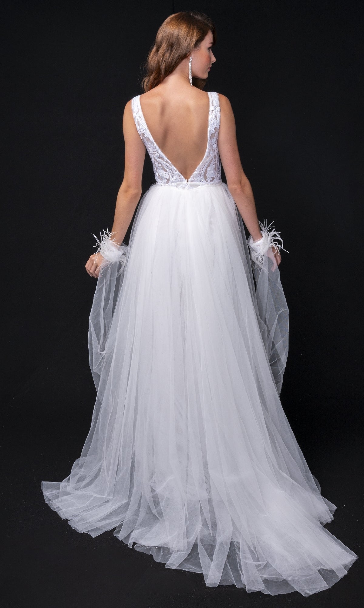 A woman in the Nina Canacci 8224 long formal dress, featuring a sleeveless design, deep V-back, and tulle skirt, stands against a black background holding the sheer fabric of her gown in both hands.