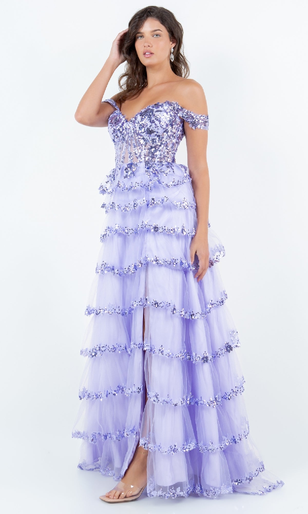 A woman wears the Ruffle Prom Ball Gown: Cinderella Couture 8228J in lavender, showcasing off-the-shoulder sequin embellishments and sheer ruffled tiers, styled with clear heels against a plain white background.