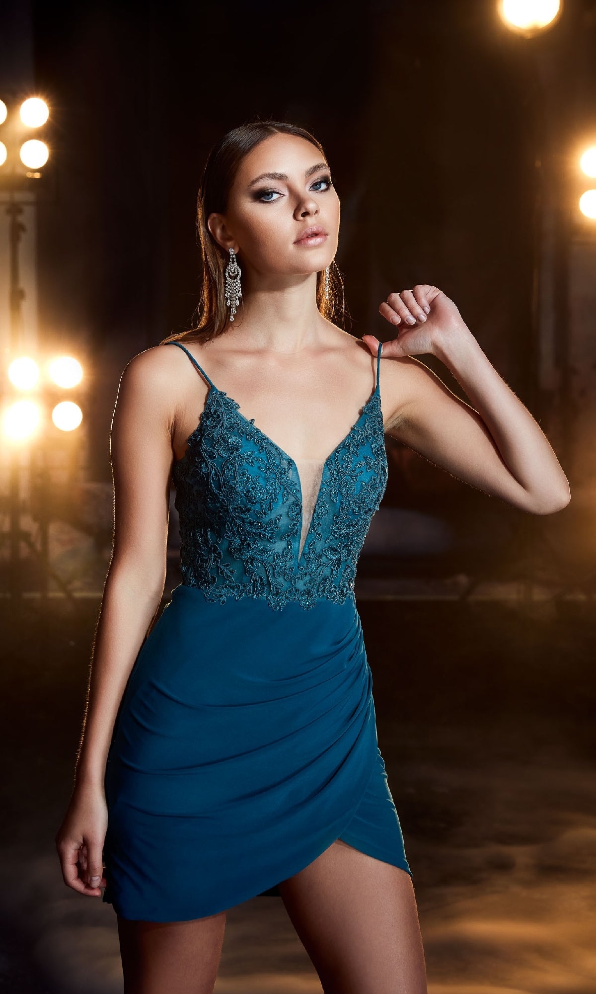 A woman in the Lace-Up Short Homecoming Dress: Aerie Couture 823, a teal sleeveless dress with lace detailing and beaded embroidery, stands before bright studio lights.