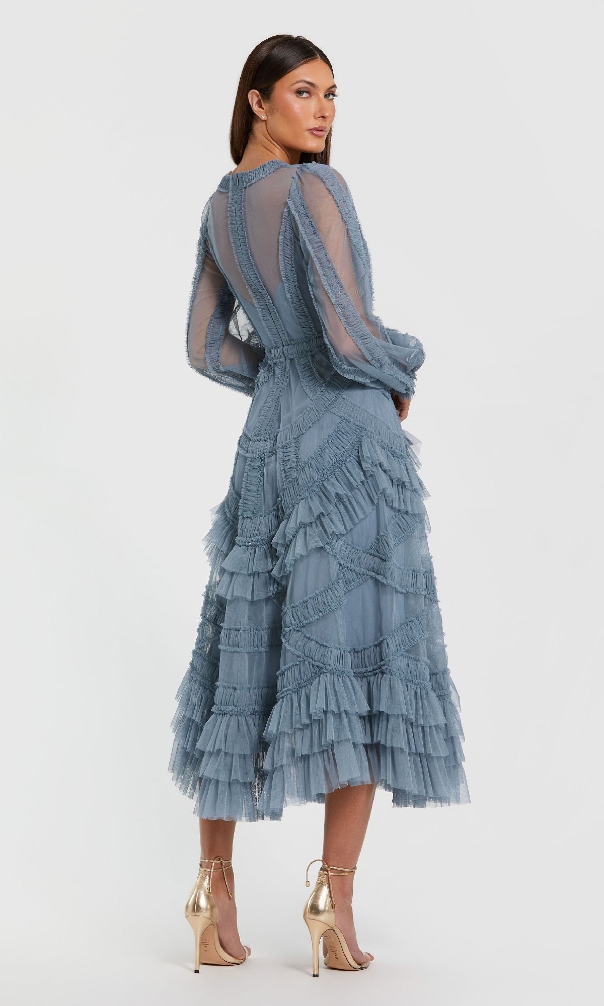 Woman wearing the Blue Midi Wedding Guest Dress: Mac Duggal 8247—a sheer, long-sleeved blue midi dress with ruffled tiered layers—stands in profile, styled with metallic high-heeled sandals for an elegant occasion-ready look.