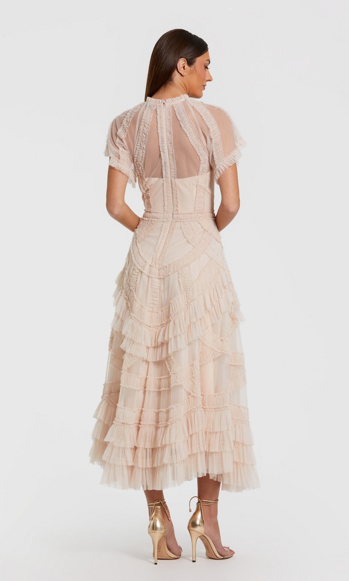A woman wearing the Mac Duggal 8249 Tea-Length Party Dress in light beige, featuring layered ruffles and short sleeves, paired with gold high-heeled sandals, stands against a plain white background facing away from the camera.