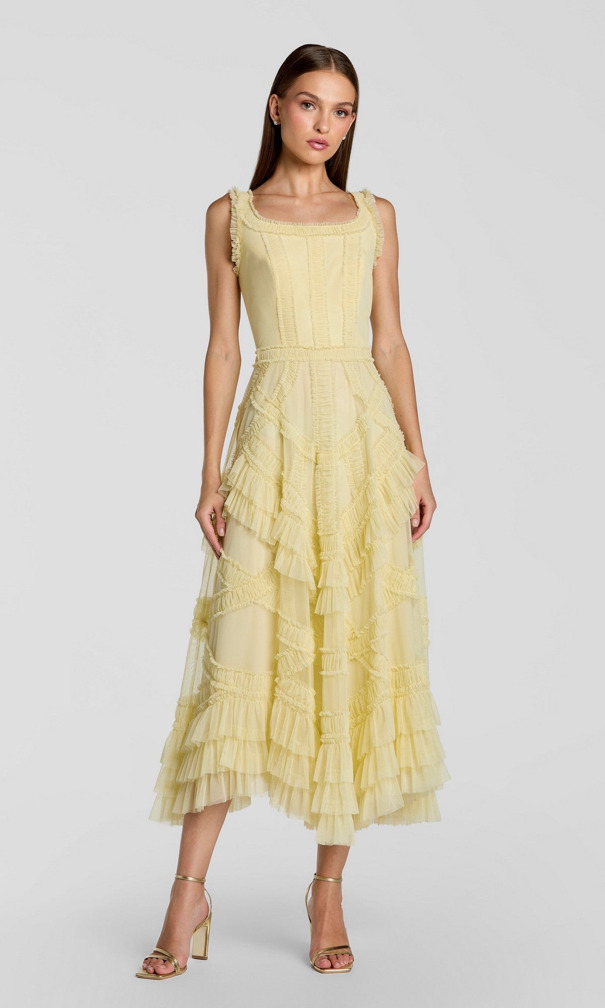 A woman wears the Mac Duggal 8250 midi wedding guest dress in buttercup, featuring sleeveless styling and micro ruffle details, paired with strappy high-heeled sandals against a plain light grey background.