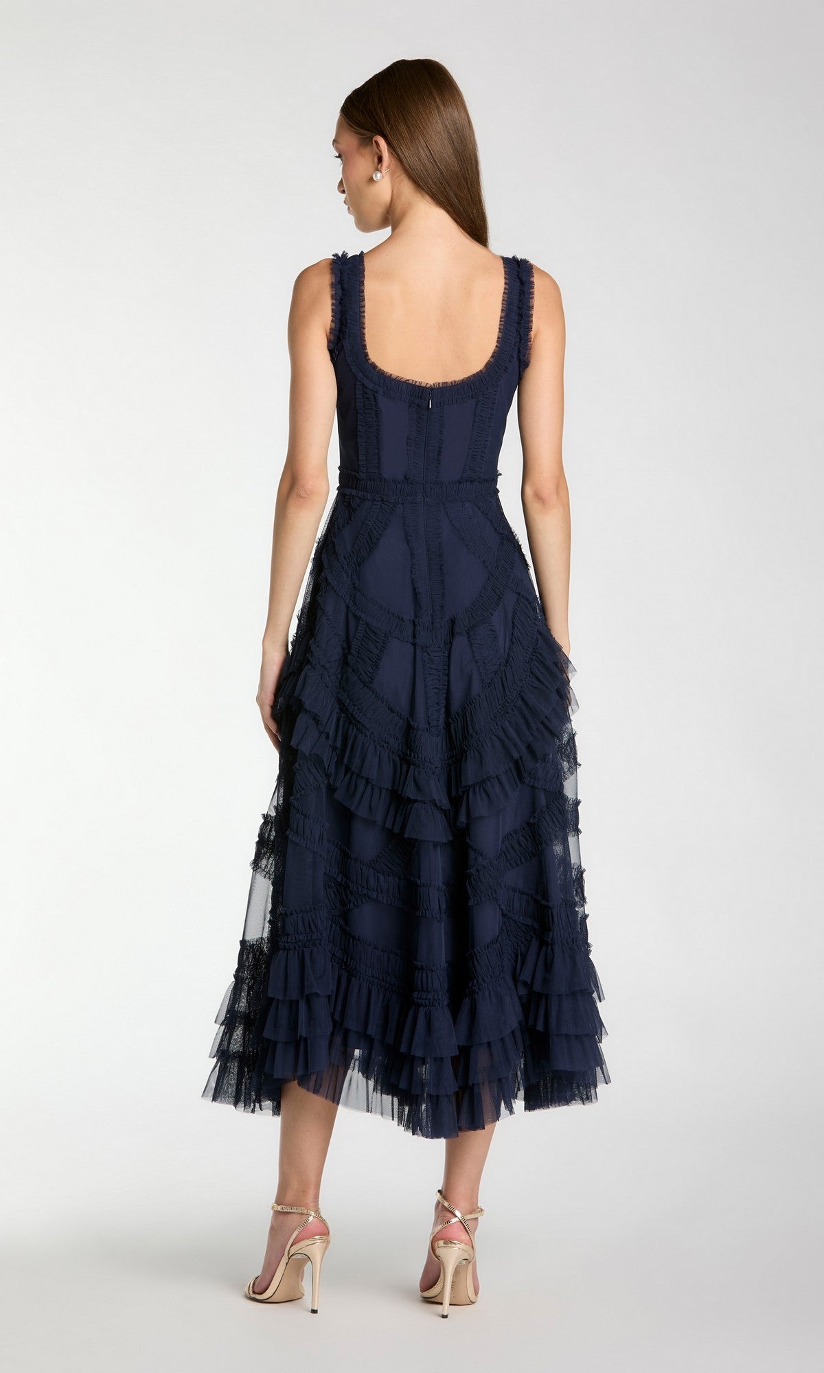 A woman stands facing away, wearing the Mac Duggal 8250 Ruffled Midi Wedding Guest Dress in navy blue with beige high-heeled sandals, set against a plain light background.
