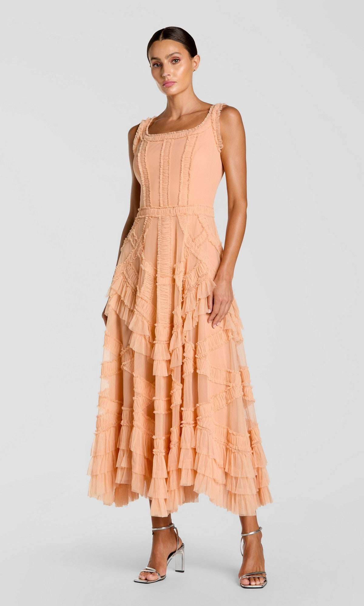 A woman in a peach Mac Duggal 8250 midi wedding guest dress with micro ruffle details and silver high-heeled sandals stands against a plain light background.